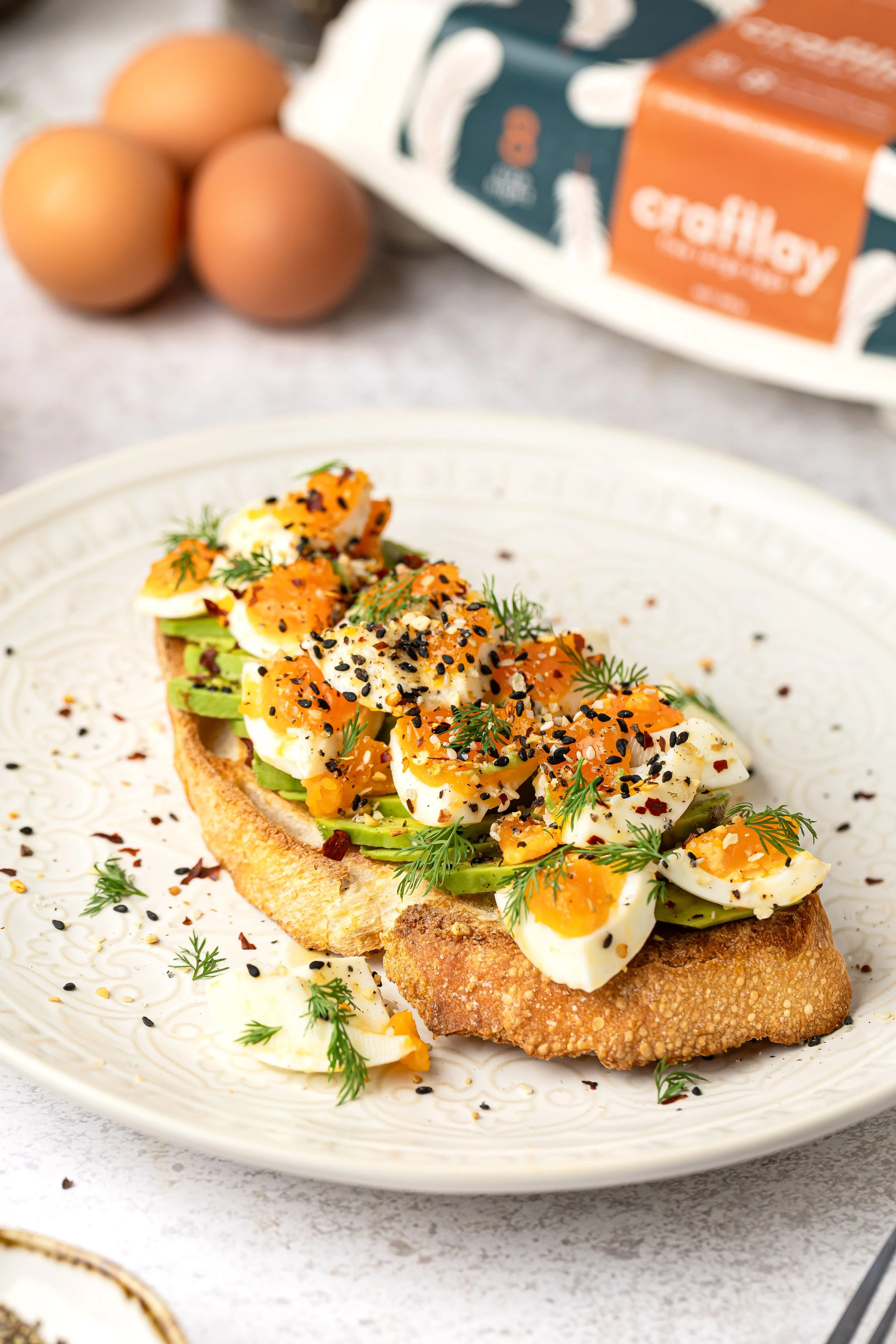 Open-faced slice of bread topped with avocado, chopped eggs, herbs, black sesame seeds, and red pepper flakes, garnished with fresh dill on a decorative plate.