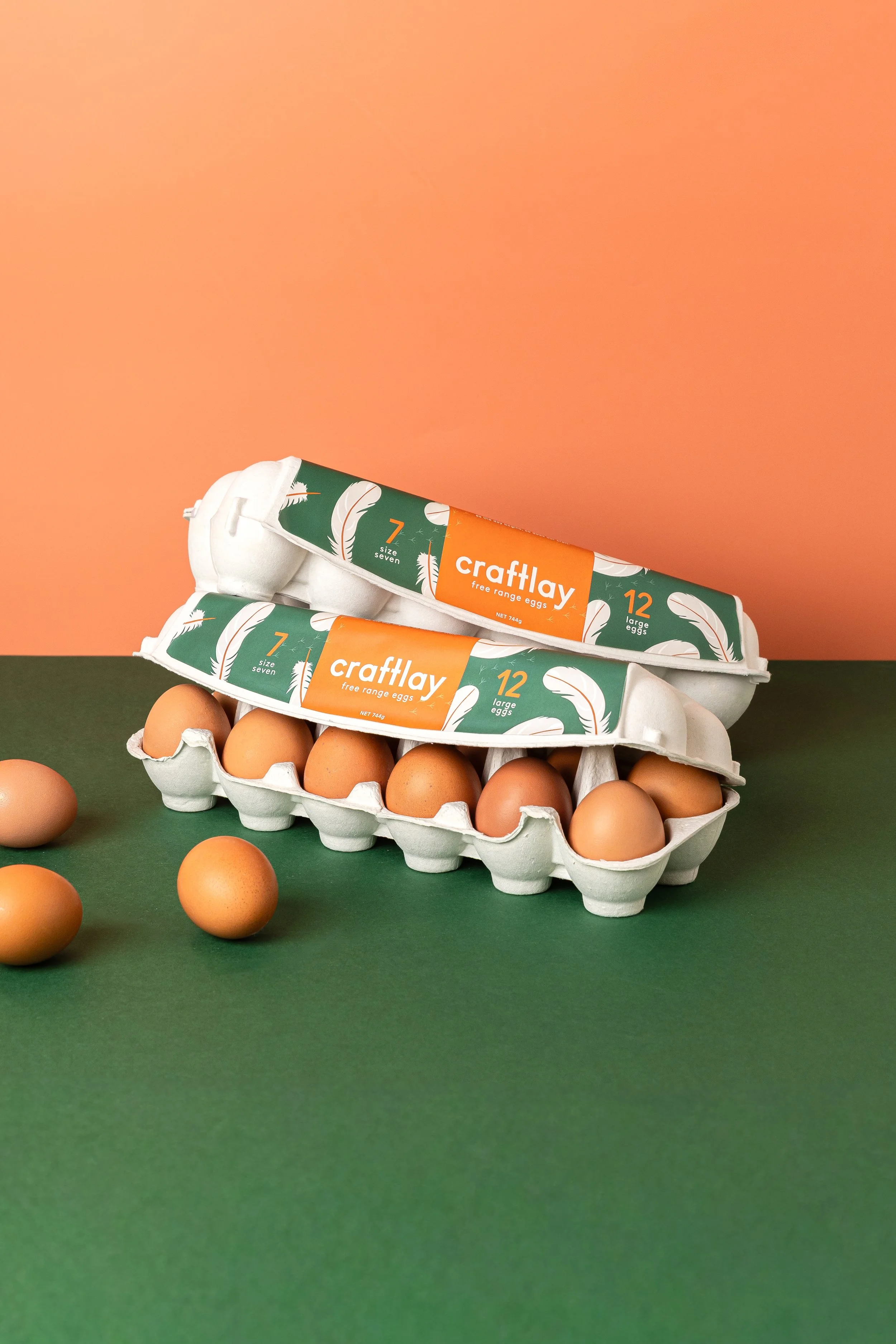 Two cartons of Craftlay free-range eggs, with a dozen eggs each, positioned on a green surface against a two-tone pink and orange background, with several eggs scattered around.
