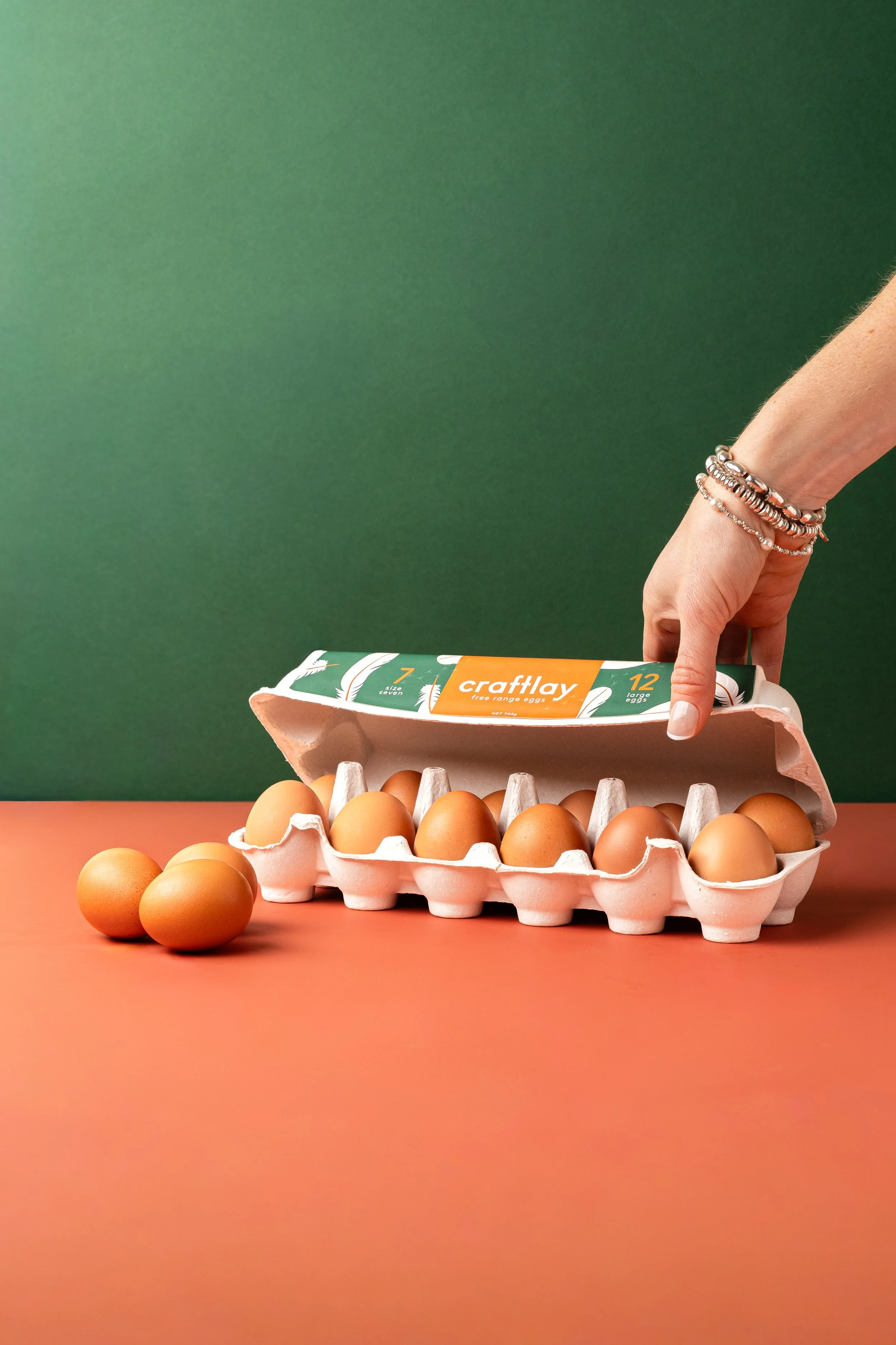 A hand with silver rings on a wrist placing a carton of 12 large eggs on a table. The carton is open with some eggs outside the carton. The background is a green wall and the table top is orange.