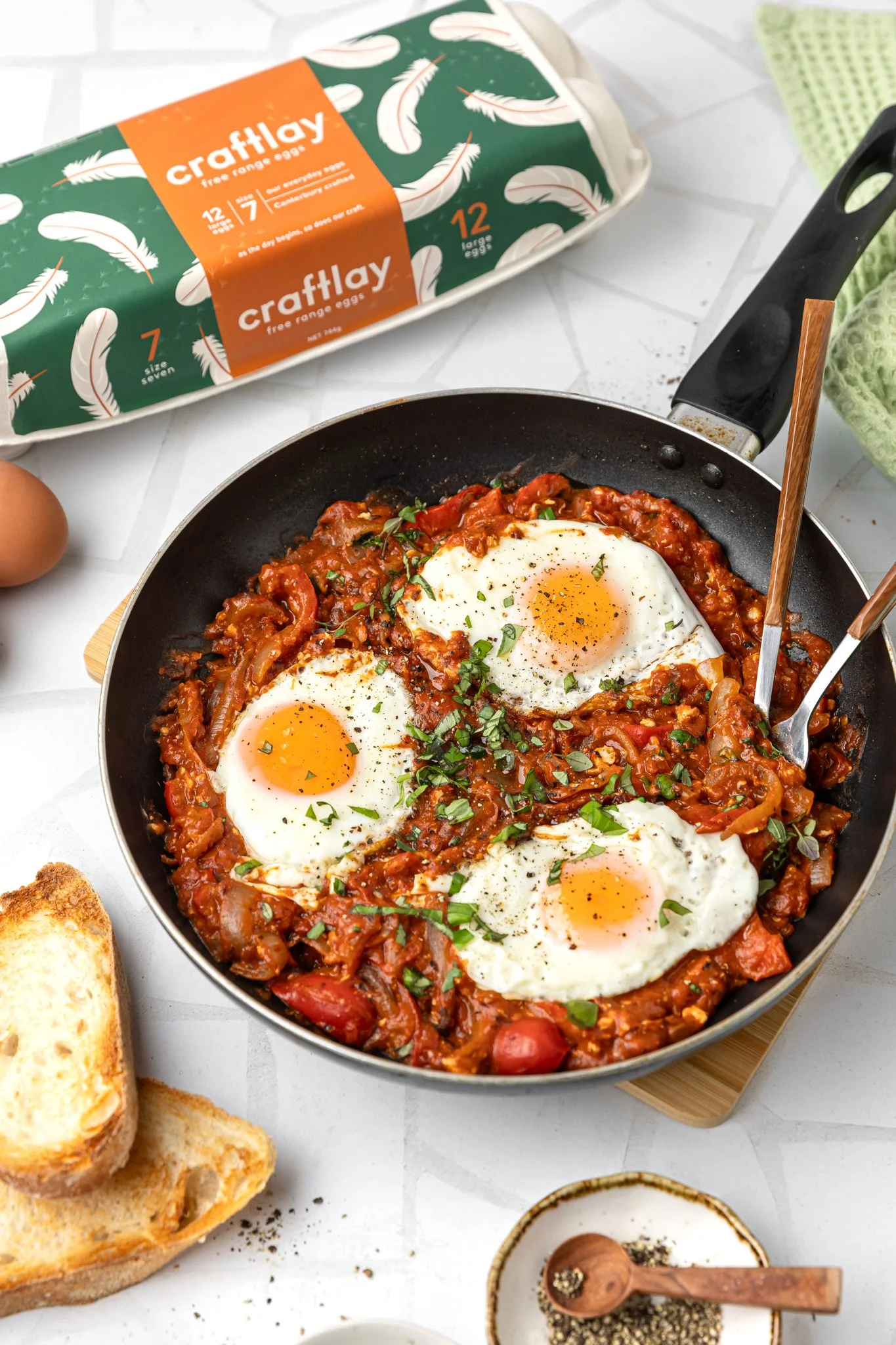 A skillet with a baked dish topped with three fried eggs, garnished with chopped herbs. Surrounding items include slices of toasted bread, a carton of eggs, and small bowls of black pepper and salt.
