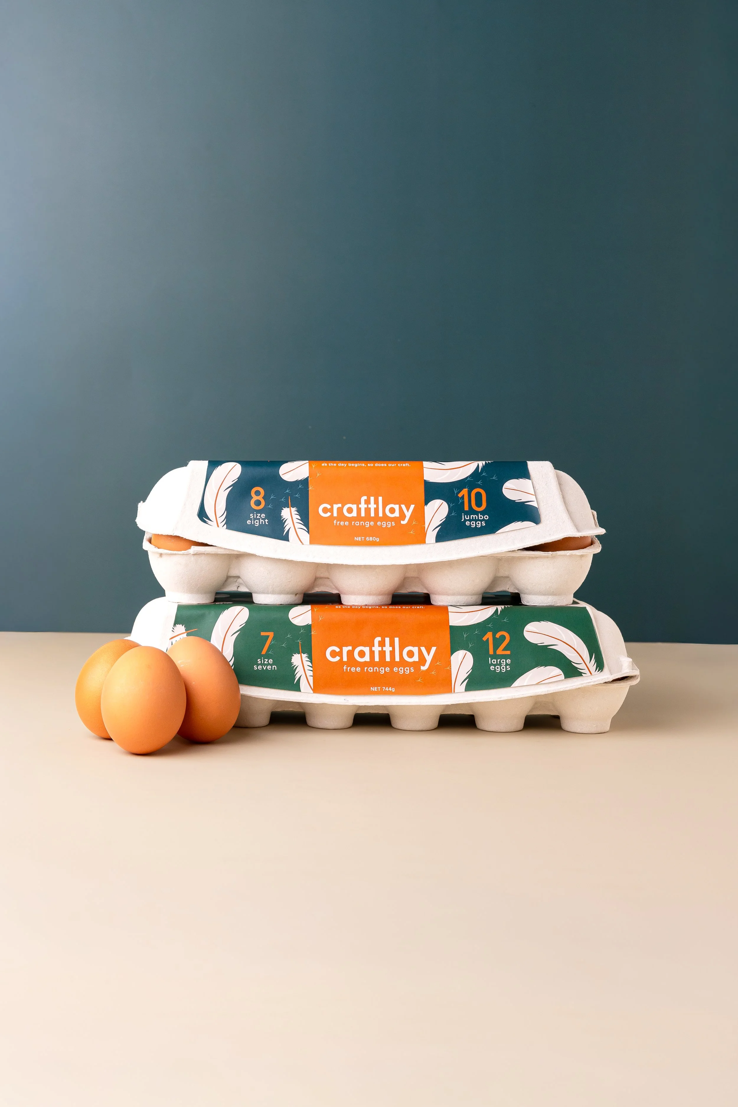 Stacked cartons of Craftlay eggs, with three hens eggs in front of the lower carton on a light surface against a dark background.