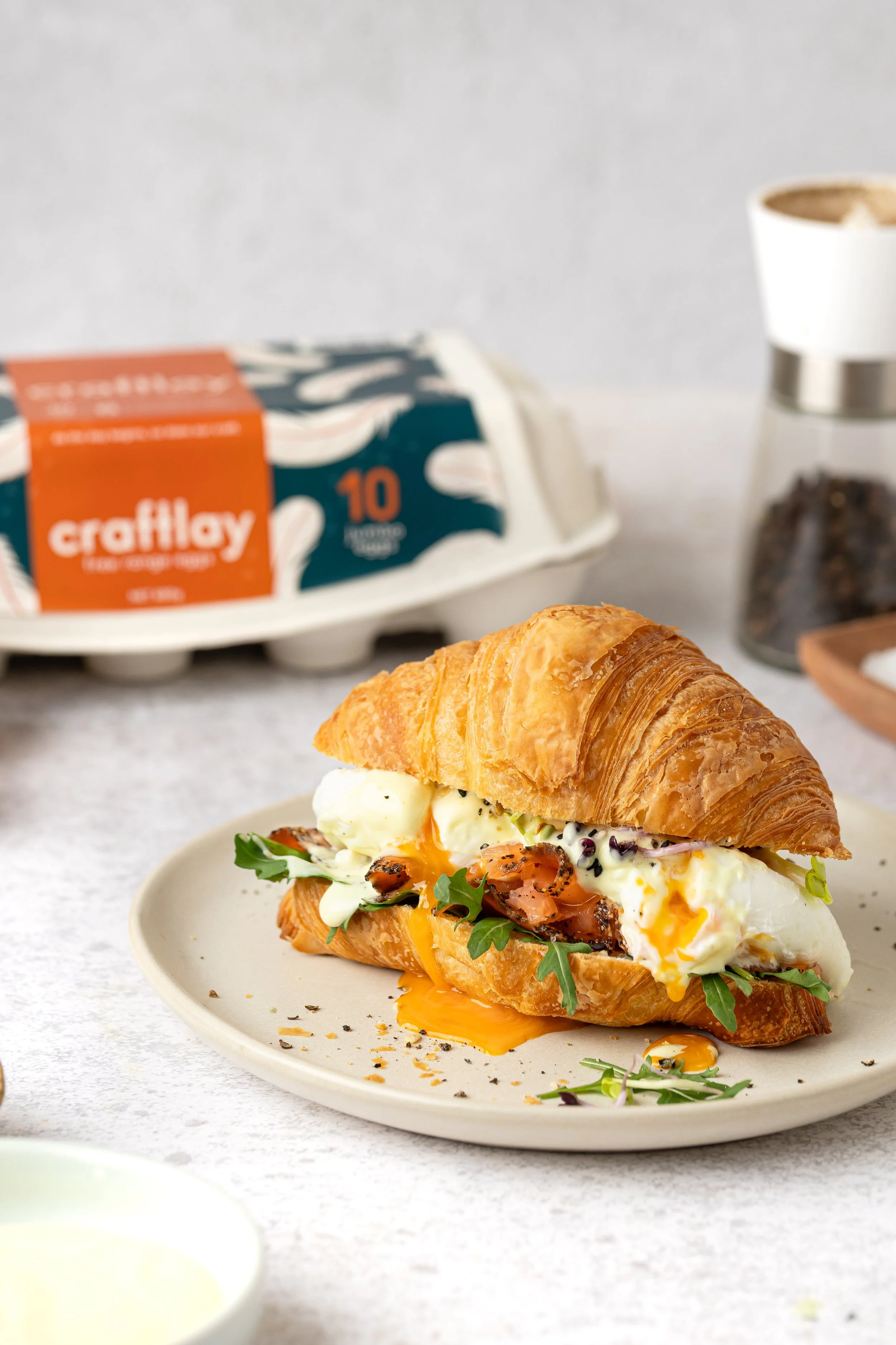 A plate with a croissant sandwich containing eggs, smoked salmon, arugula, and condiments. In the background, there is a carton of 10 eggs, a cup of coffee, and a container of coffee grounds.