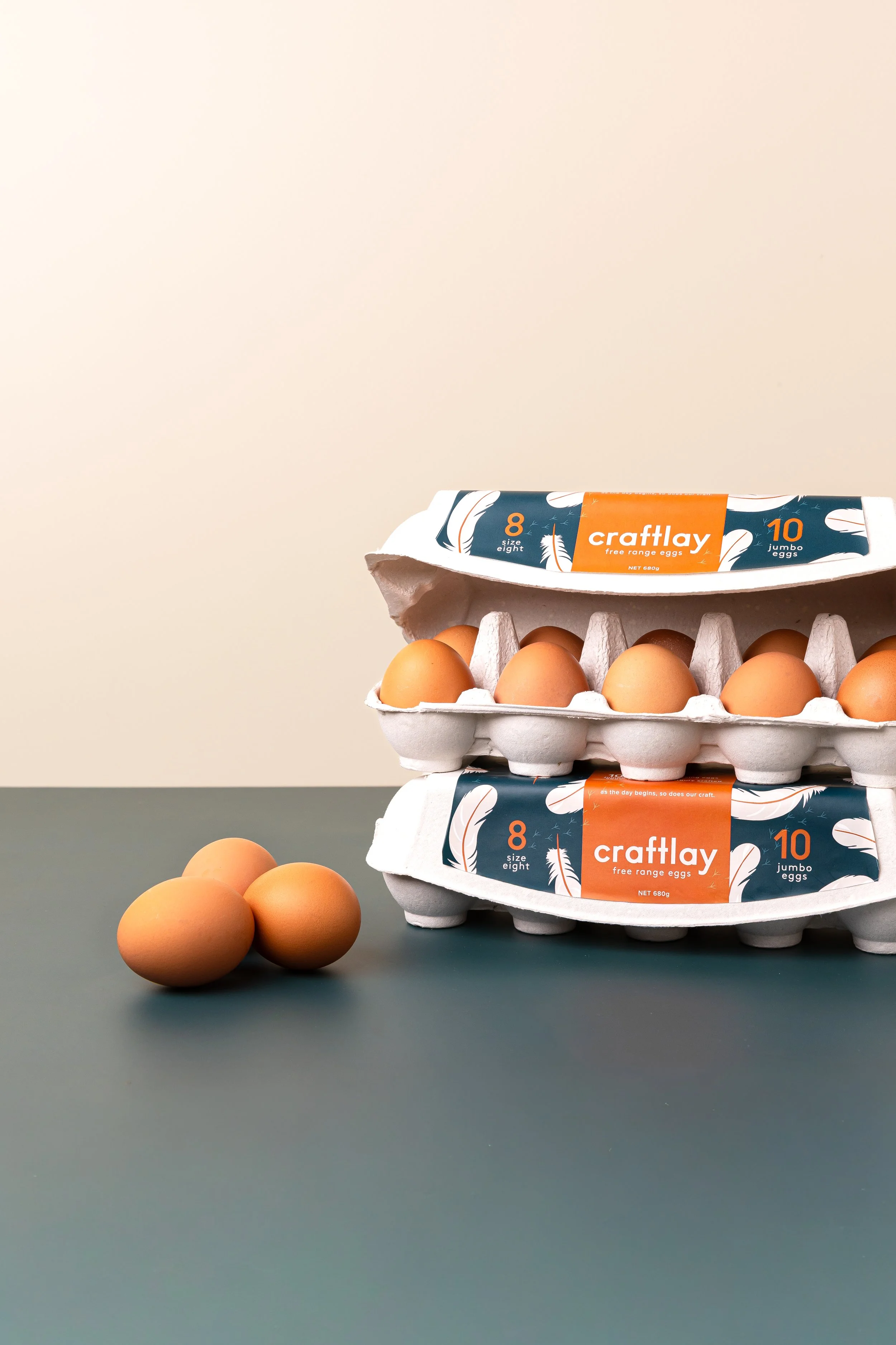 Two cartons of Craftlay free-range eggs stacked on a dark gray surface, with brown eggs in the cartons and some brown eggs outside the cartons on the surface. The packaging indicates the eggs are size eight and jumbo size ten.