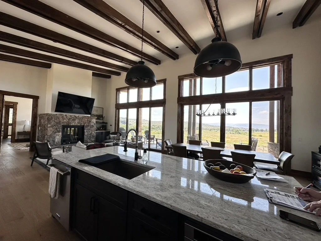 Open-concept living and dining area with a kitchen island, large windows showing a scenic outdoor landscape, a stone fireplace, a mounted flat-screen TV, and modern pendant lighting.