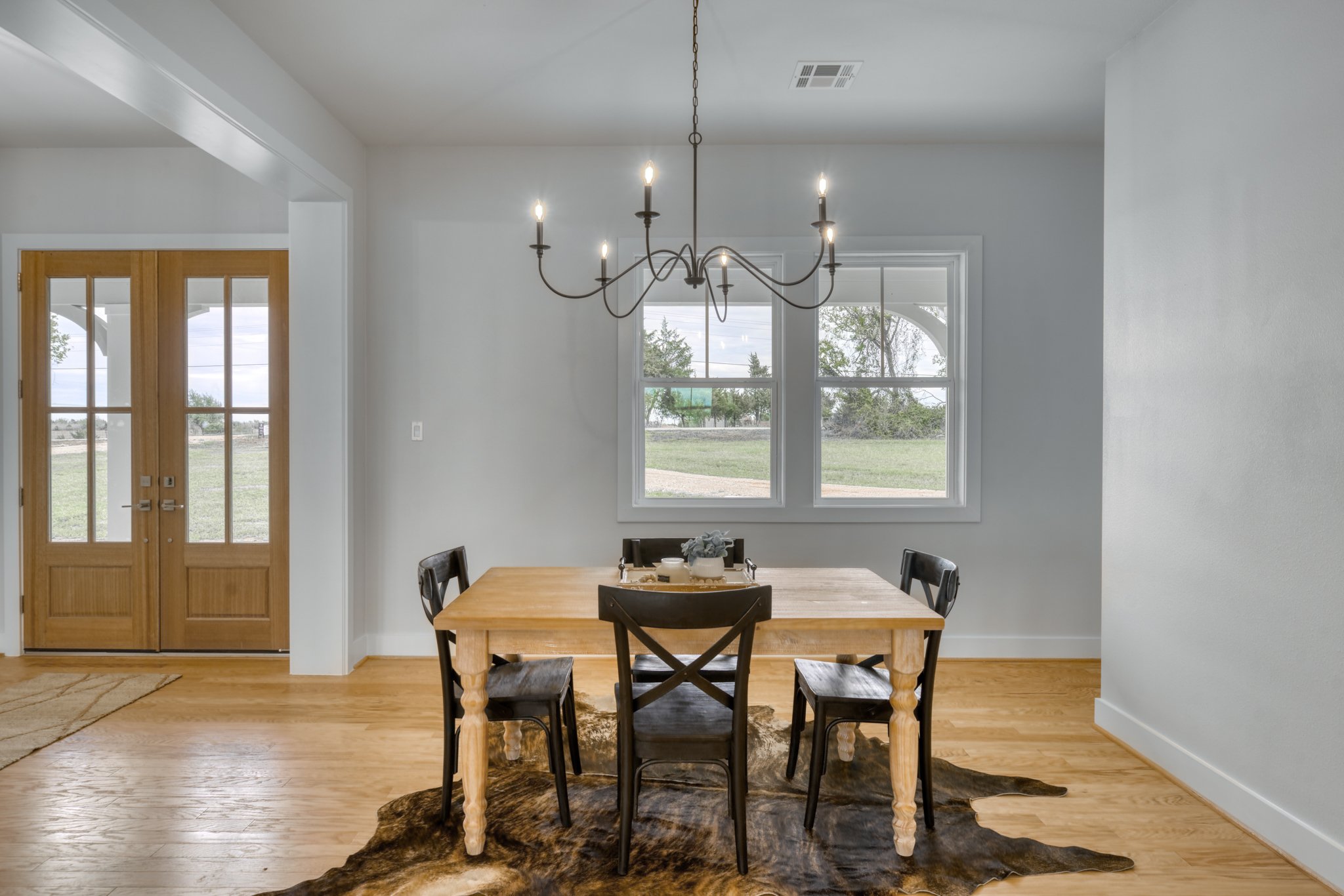 Dining room with a wooden table and five black chairs, a black chandelier hanging from ceiling, large window showing outdoor landscape, double doors with glass panels, and a cowhide rug on hardwood floor.