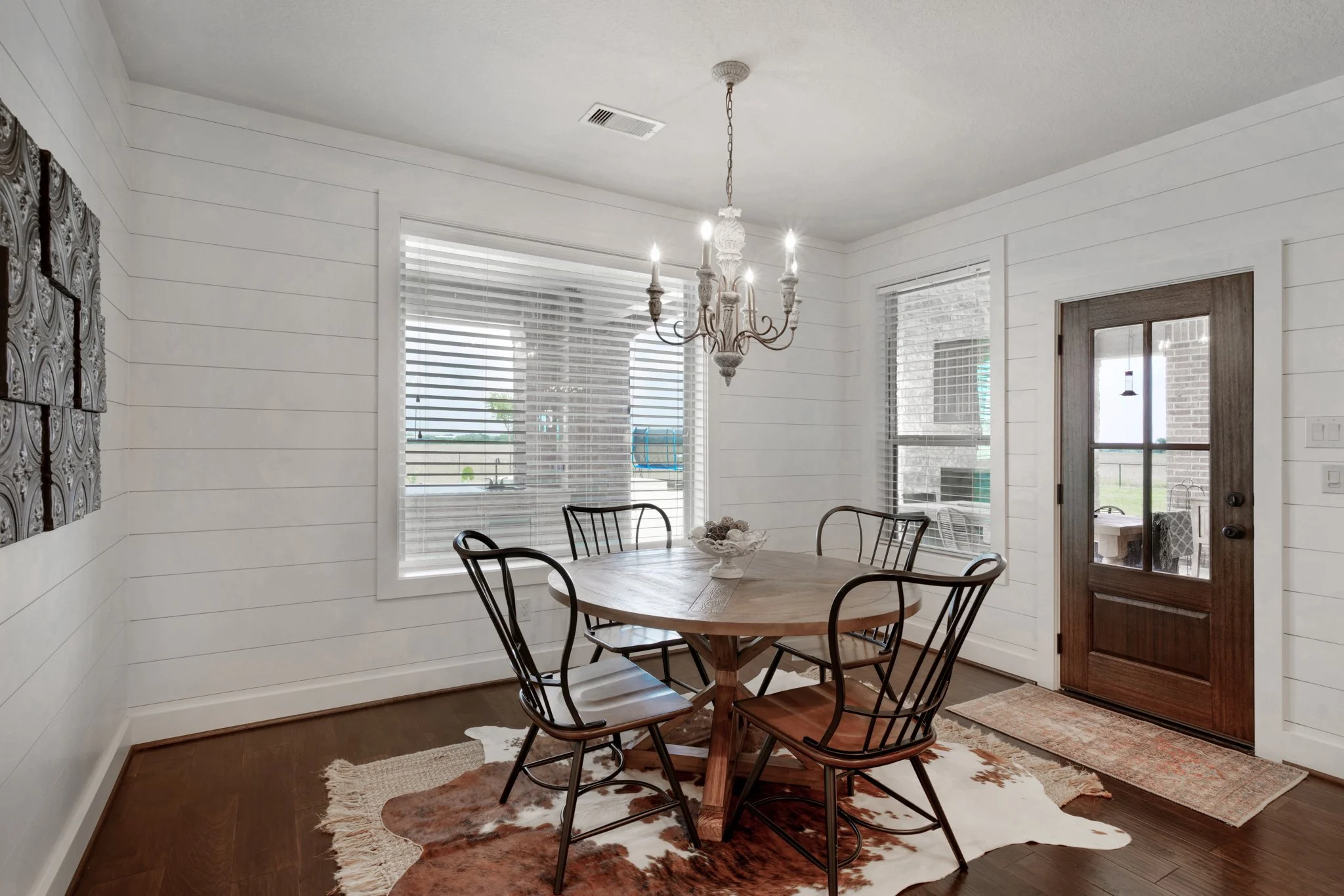 A dining room with white shiplap walls, a wooden table with five chairs, a chandelier, and a cowhide rug. There are large windows with blinds and a glass door leading outside.
