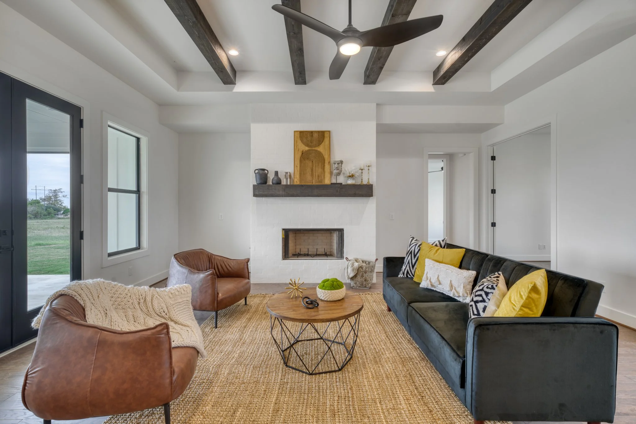 Modern living room with white walls, a white brick fireplace, and dark wooden ceiling beams. The room has large windows and a glass door leading outside. There is a black velvet sofa with yellow and black-and-white pillows, two brown leather armchairs with a knitted throw, and a round wooden coffee table on a natural fiber rug. Above the fireplace are decorative items including vases, sculptures, and framed artwork.