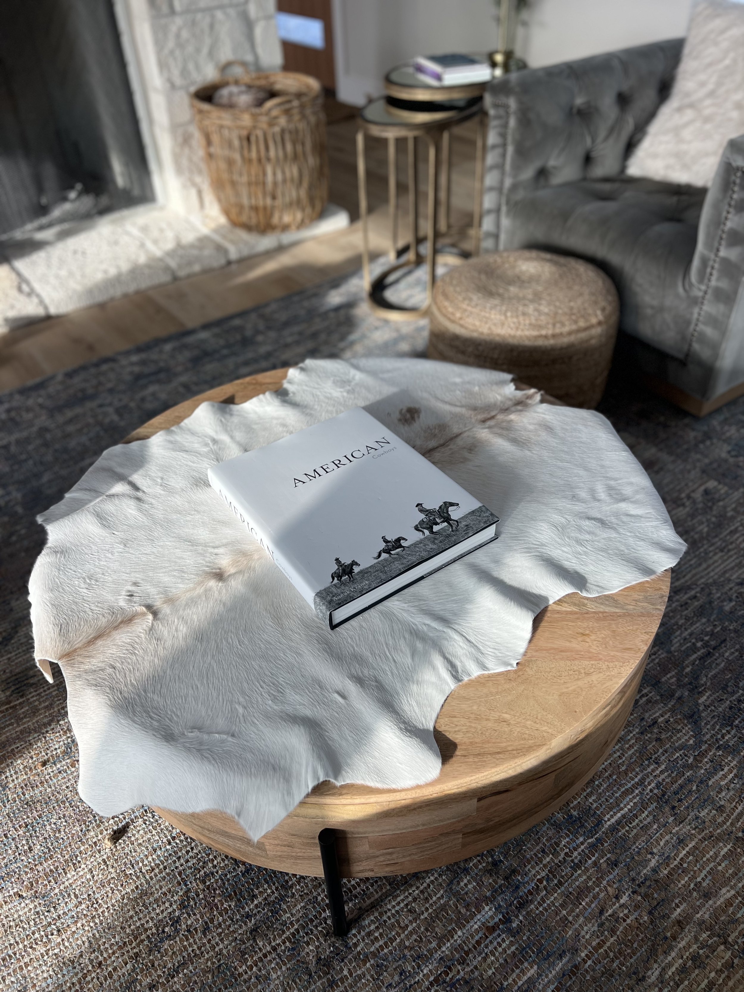 A round wooden coffee table with a white leather or faux leather animal hide cloth on top. A book titled 'American' is placed on the cloth, featuring black-and-white illustrations of a cowboy on horseback. The background includes a stone fireplace, woven basket, round side table, rattan ottoman, and a grey tufted armchair with a pillow.