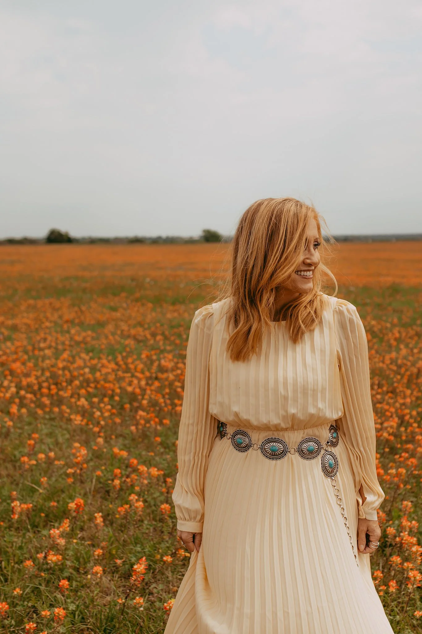 A woman with red hair wearing a cream-colored pleated dress with a decorative belt stands in a field of orange flowers, smiling and looking to the side.