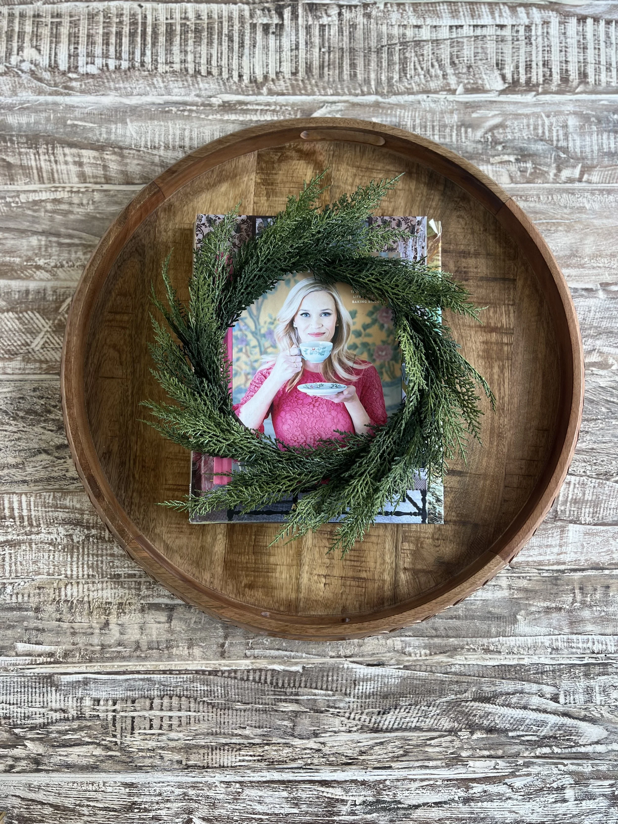 A wooden tray with a green Christmas wreath and a magazine featuring a woman in a red dress holding a teacup on top of a wooden table.