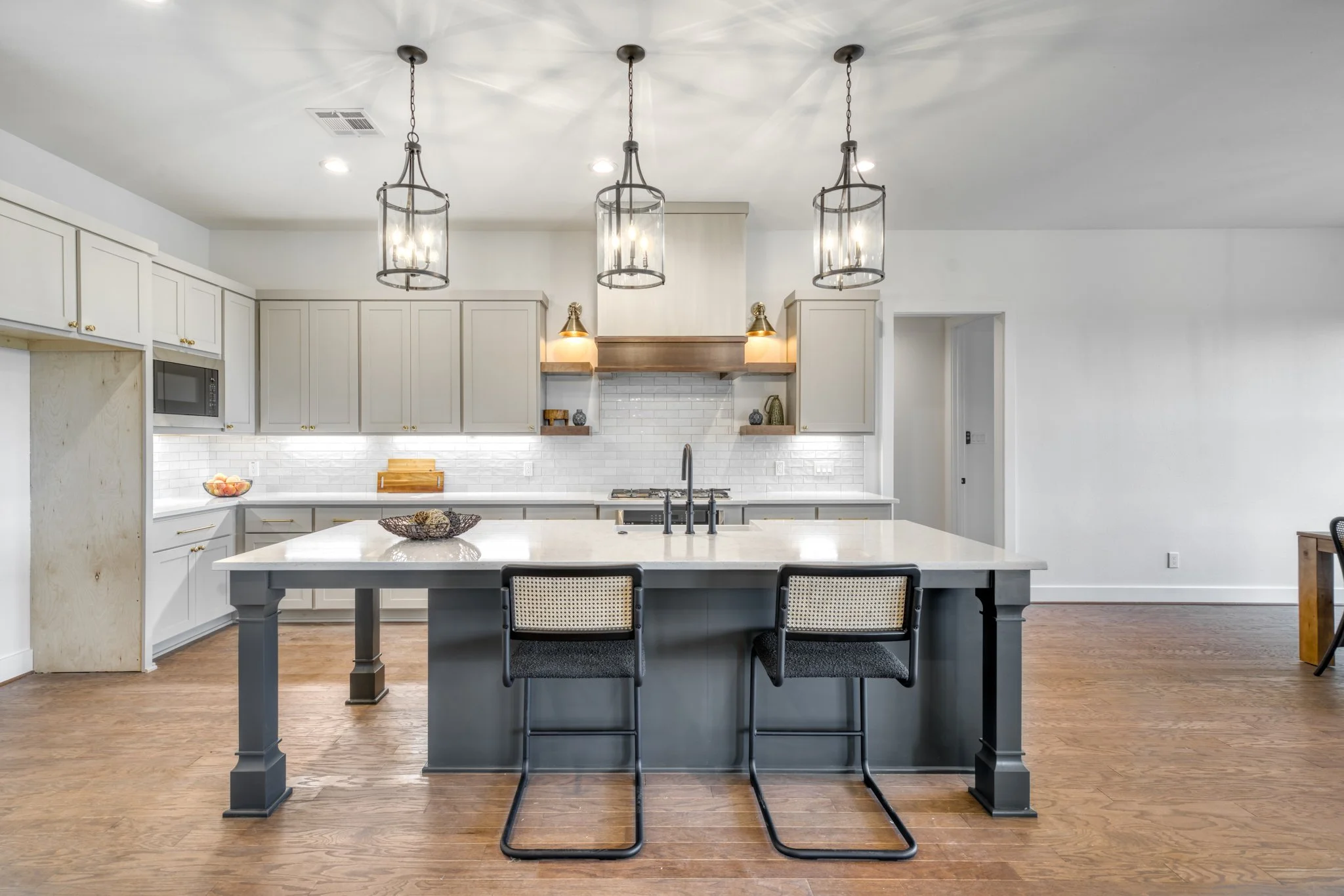 Modern kitchen with white cabinets, marble island counter, three hanging pendant lights, and two black barstools with woven seats.