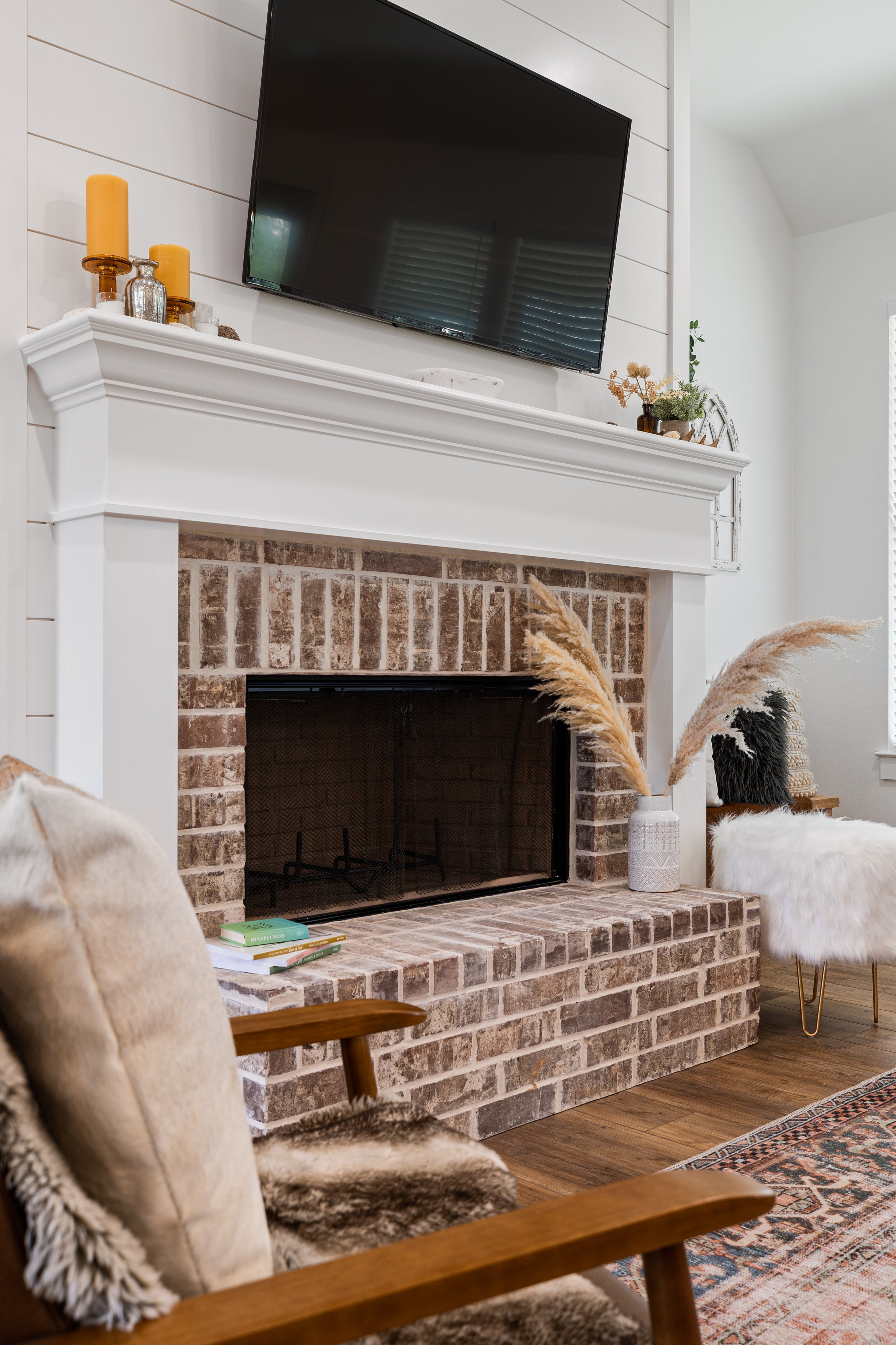 Living room with brick fireplace, large flat-screen TV above mantel, plush chair with throw pillow, white fur-covered chair, decorative vases, books, and plants.
