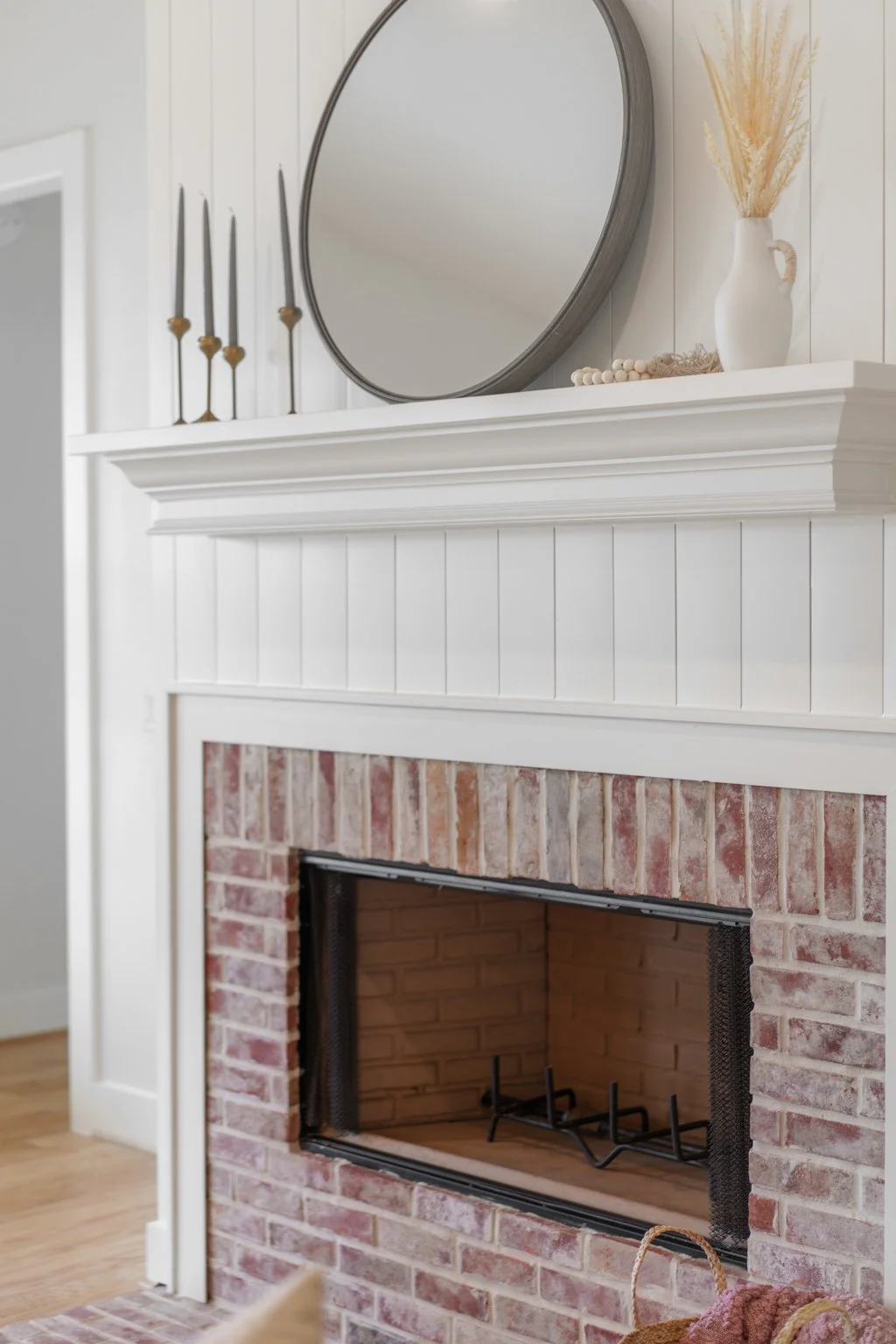 A white mantel with a round mirror, tall candle holders, and a white vase with dried plants over a brick fireplace.