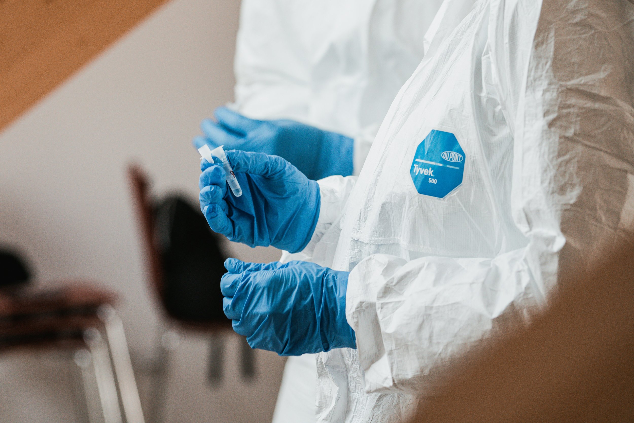 A healthcare worker in a white protective suit with a blue Tyvek logo on it is holding a syringe and preparing to administer a vaccine.