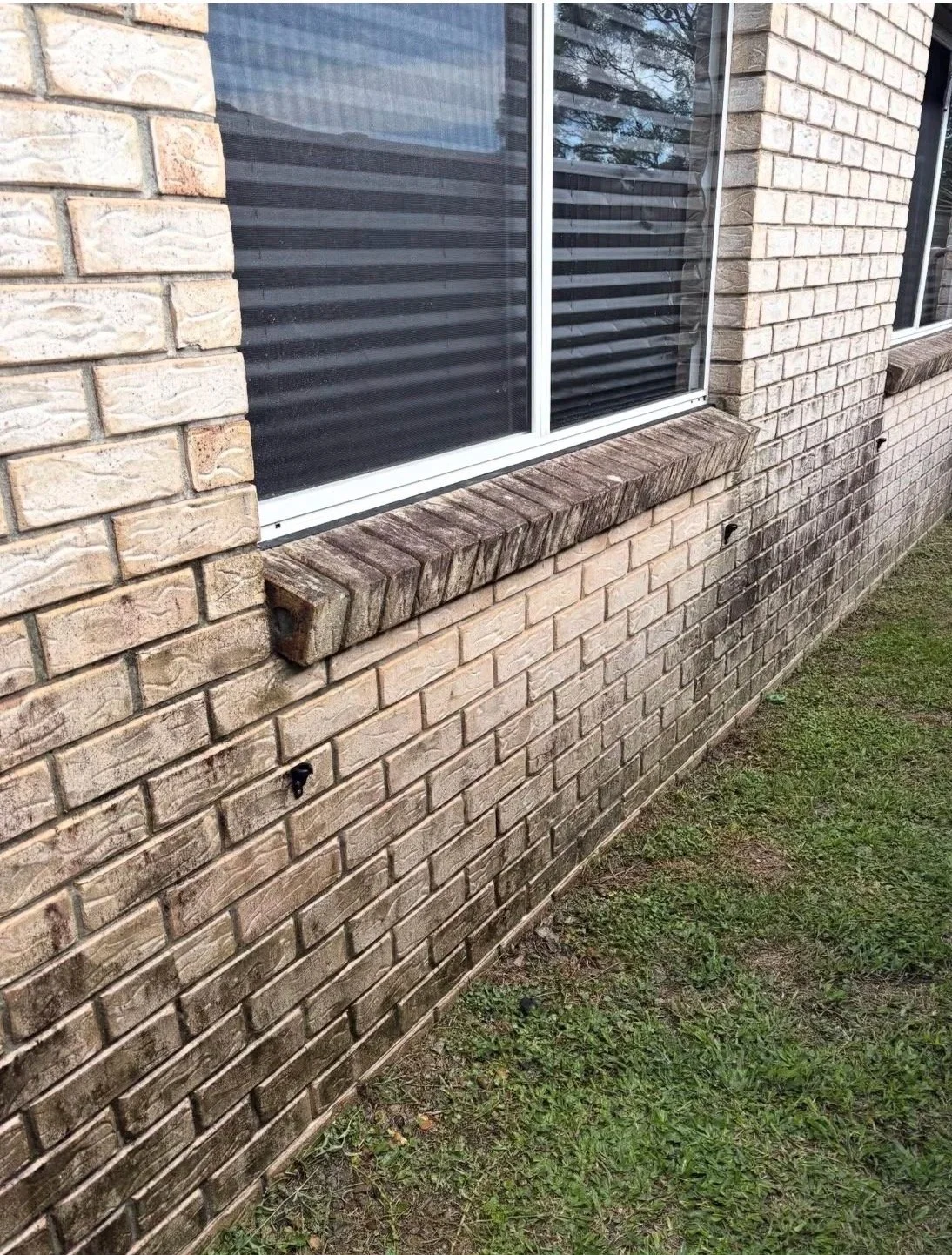 A brick house exterior with a window, and an old, warped wooden board placed underneath the window sill on the brick wall.