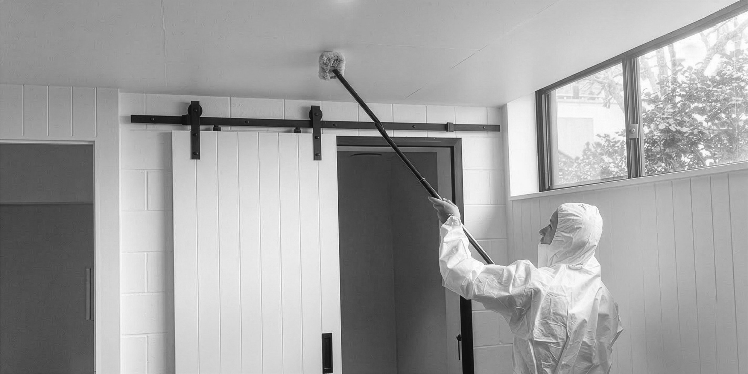Person in protective suit using a cleaning tool on a sliding barn door in a room with white walls and large windows.