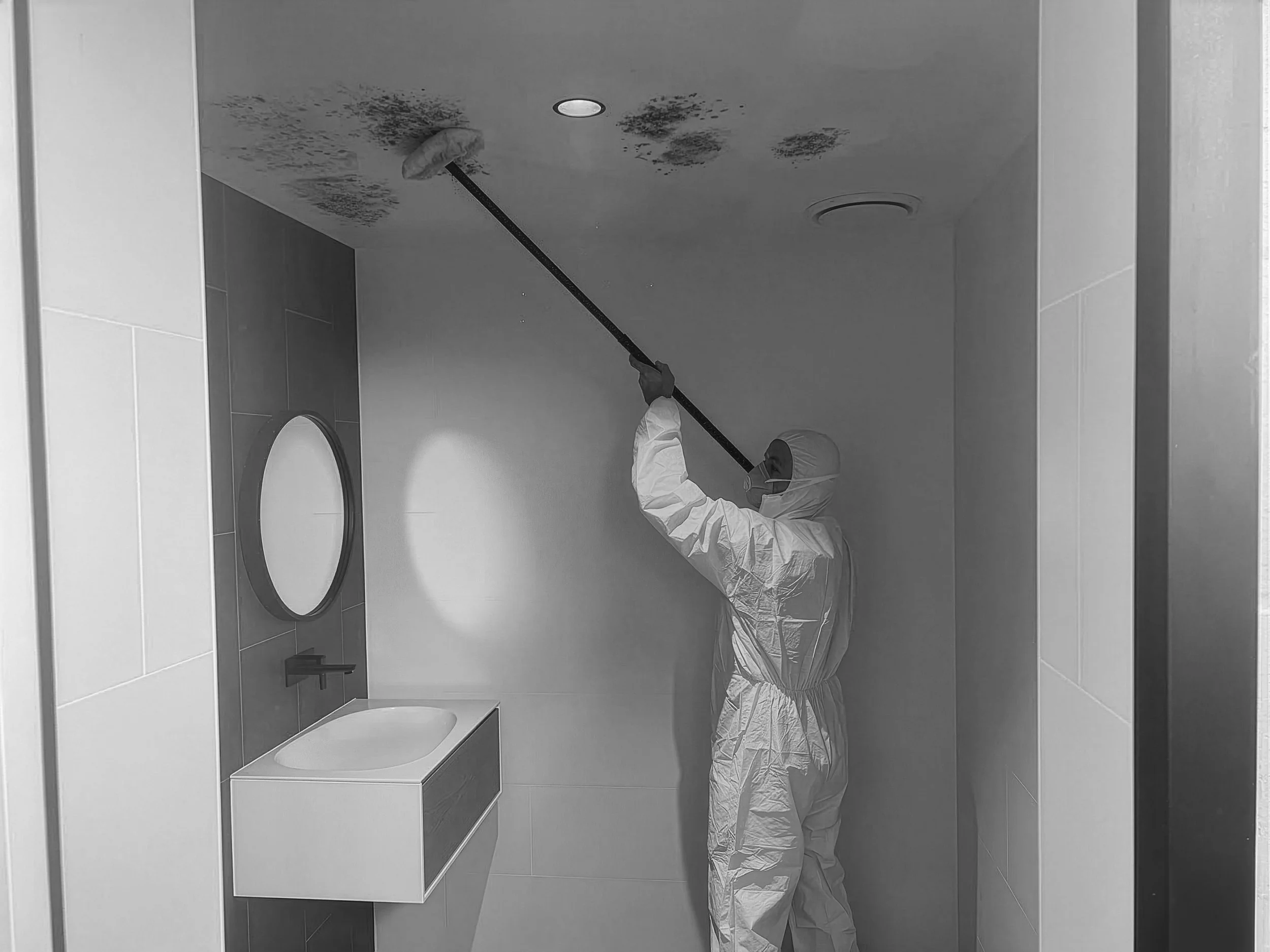 A person wearing protective suit, mask, and gloves is using a long pole to clean or inspect the ceiling of a bathroom, which has water stains or mold.