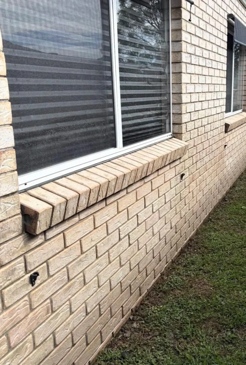 A brick exterior wall of a building with three large windows, each with a white frame and black blinds. The middle window has a small brick ledge beneath it.