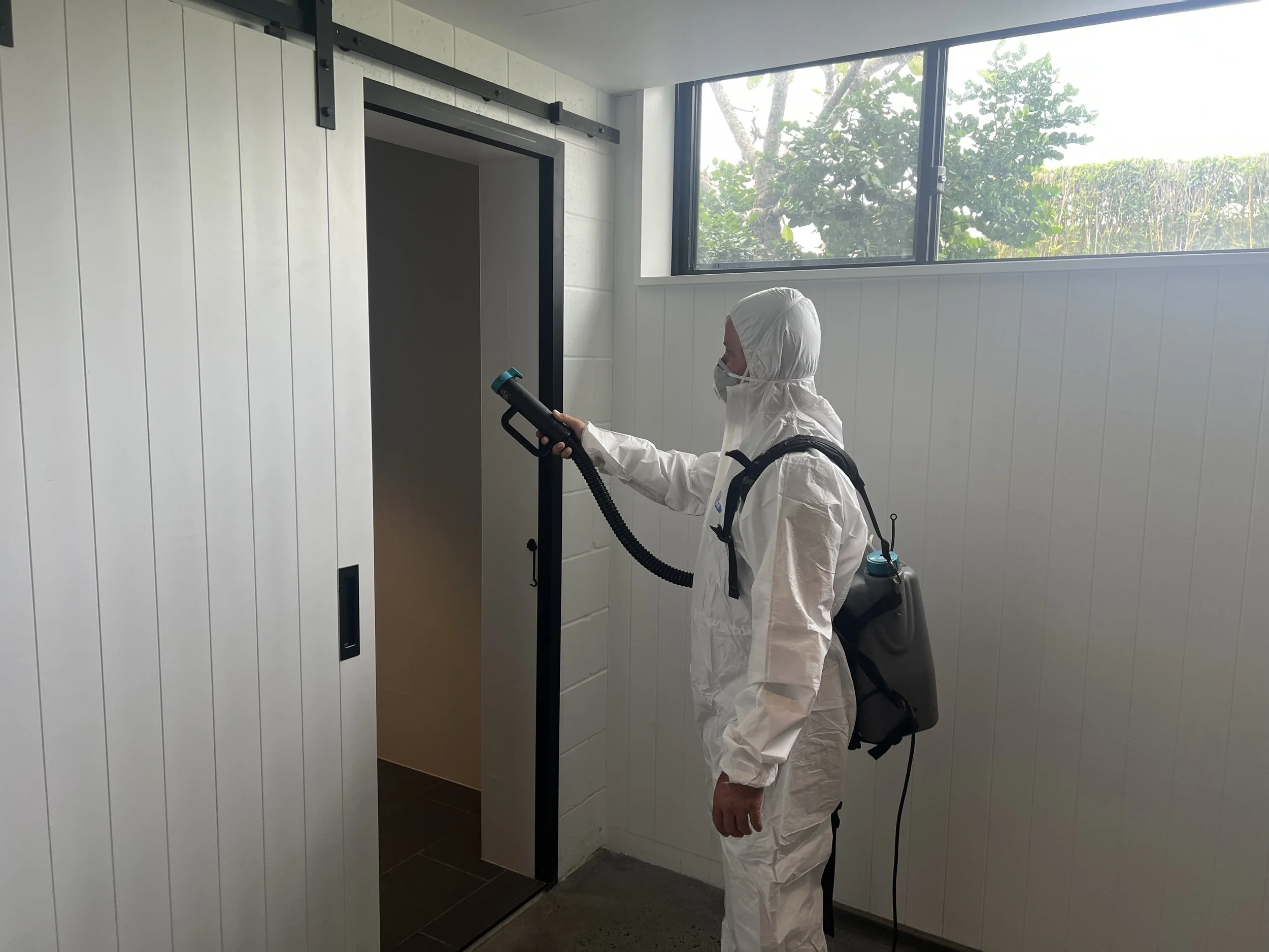 Person in white protective suit and face mask disinfecting a door frame with a handheld sprayer inside a building with white walls and large window.