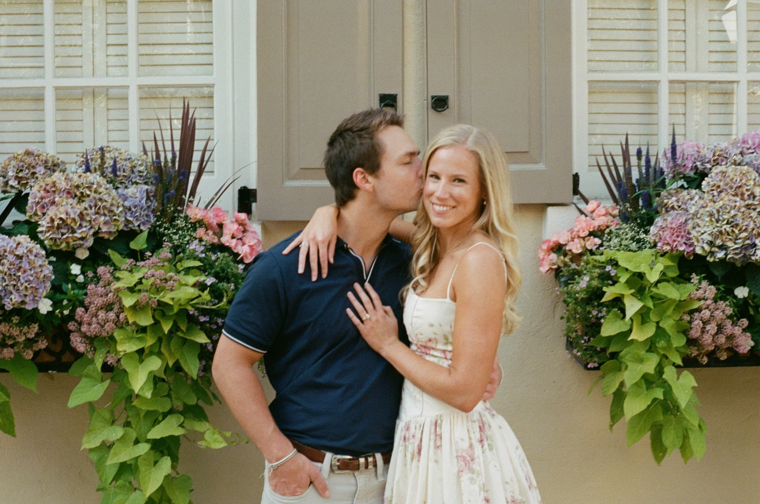 A happy couple stands close together in front of a house with beige siding and white window shutters, surrounded by abundant pink and purple hydrangea bushes, as the man kisses the woman's temple and the woman grins at the camera.