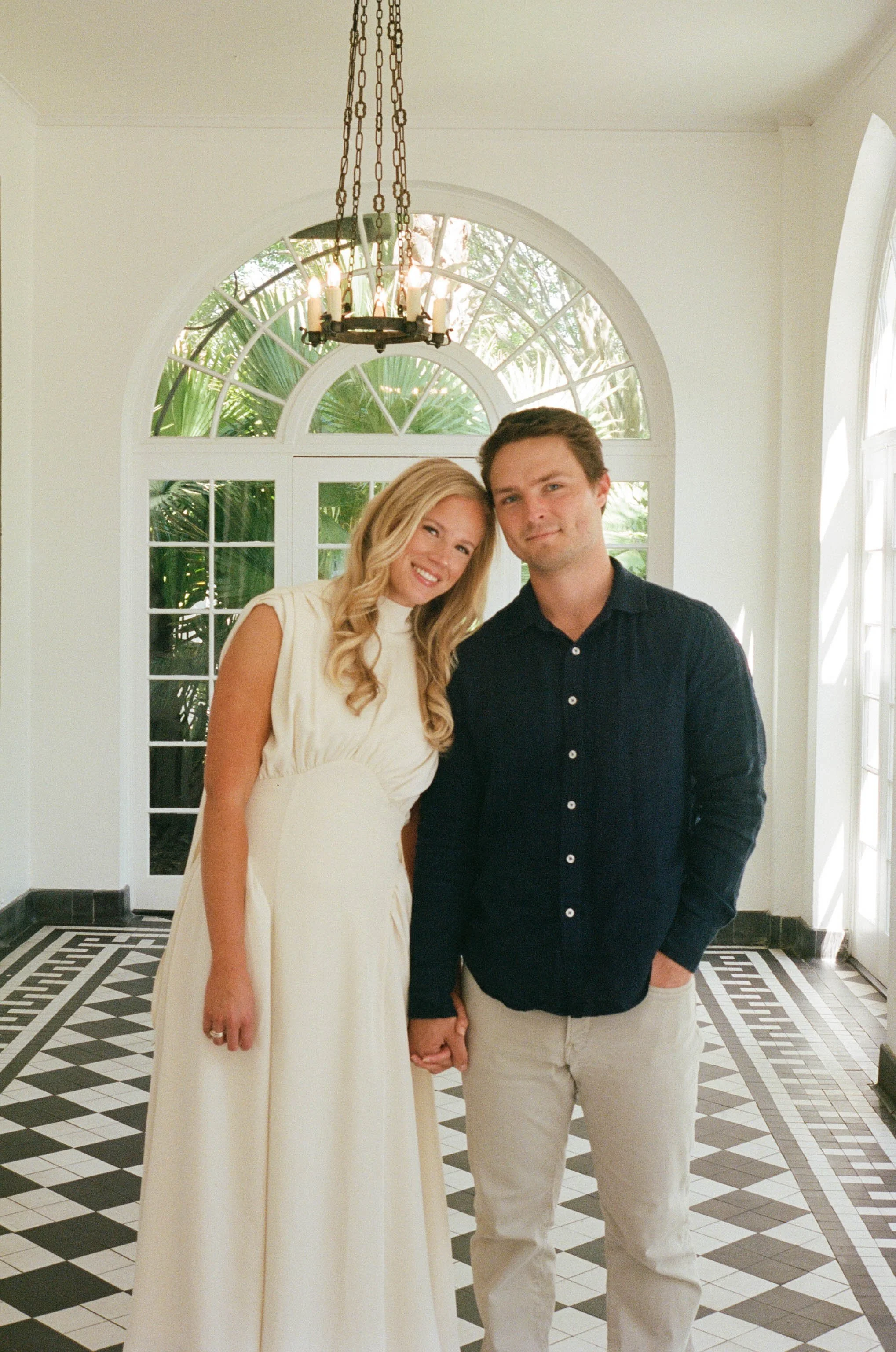 A couple standing hand in hand inside a bright room with an arched window overlooking greenery. The woman is wearing a cream-colored dress with ruffled sleeves, and the man is wearing a navy blue shirt and beige pants.