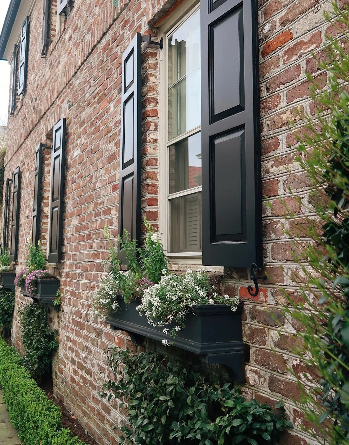 A brick house with multiple windows, each with black shutters and window boxes filled with flowers.
