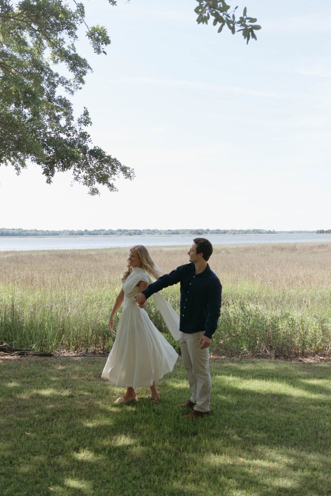 A couple standing outdoors by a lake, with the woman in a white dress and the man in a dark shirt and light pants. They are holding hands with the man leading the woman, who is smiling. There are trees overhead and tall grass near the water.