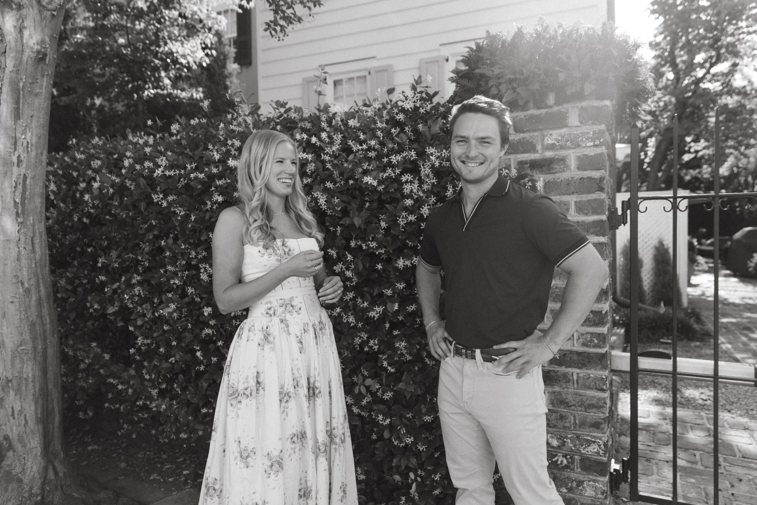 A black-and-white photo of a smiling man and woman standing outdoors near a brick wall and lush foliage, enjoying a sunny day.