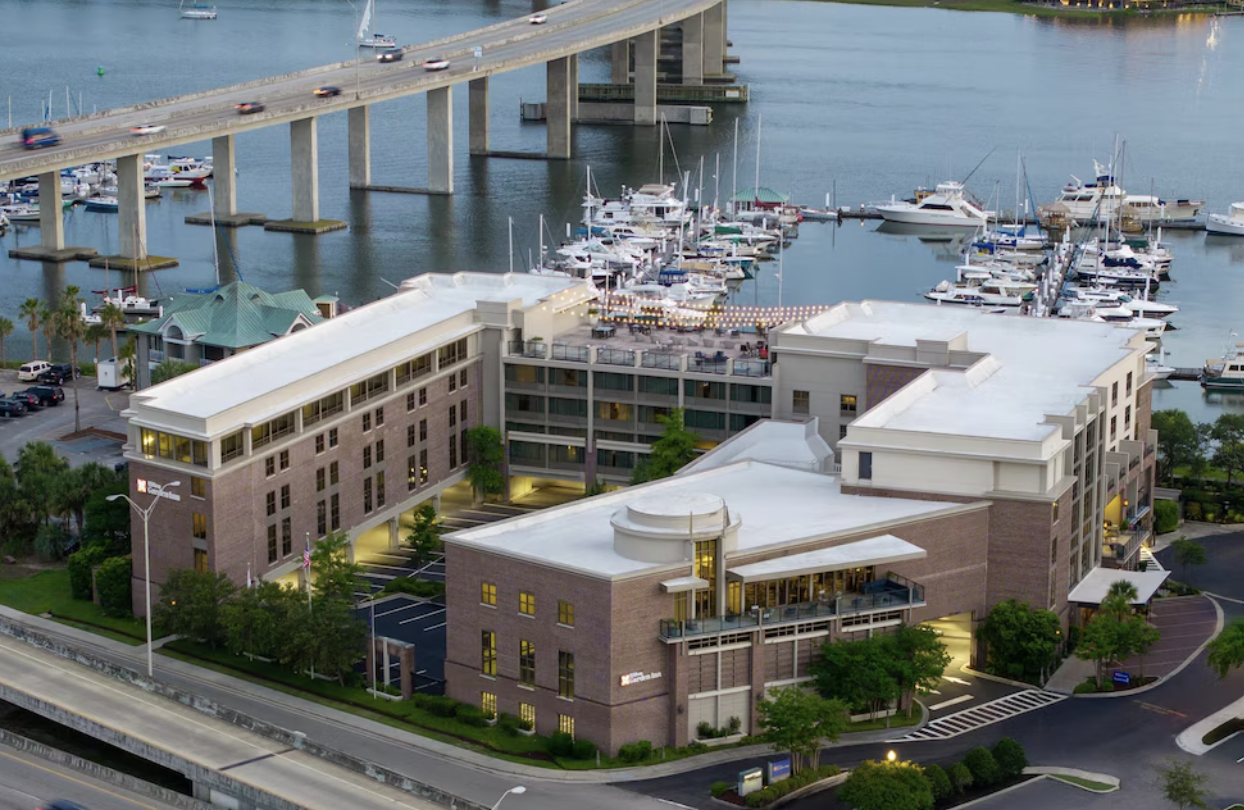 Aerial view of a marina with boats docked, a large modern hotel or apartment building, and a bridge crossing over the water in the background.