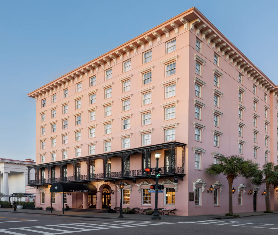 Pink multi-story hotel with a black balcony and awning, situated on a street corner with traffic lights and palm trees.