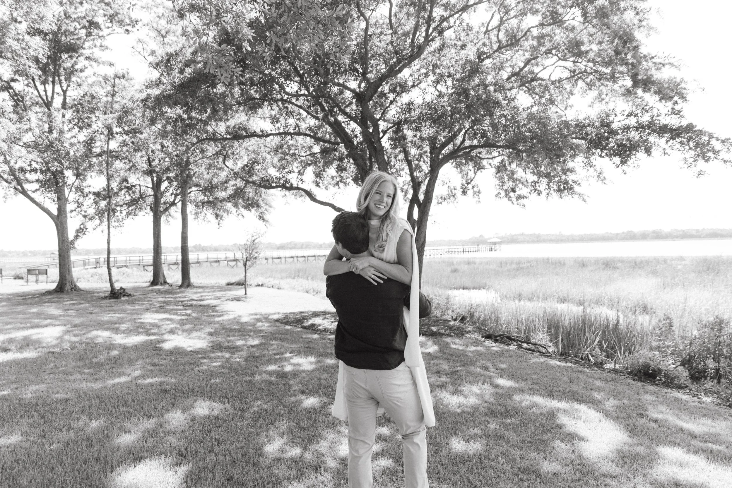 A couple sharing a hug outdoors on a sunny day, with a woman smiling and a man holding her from behind in front of a large tree and a body of water.