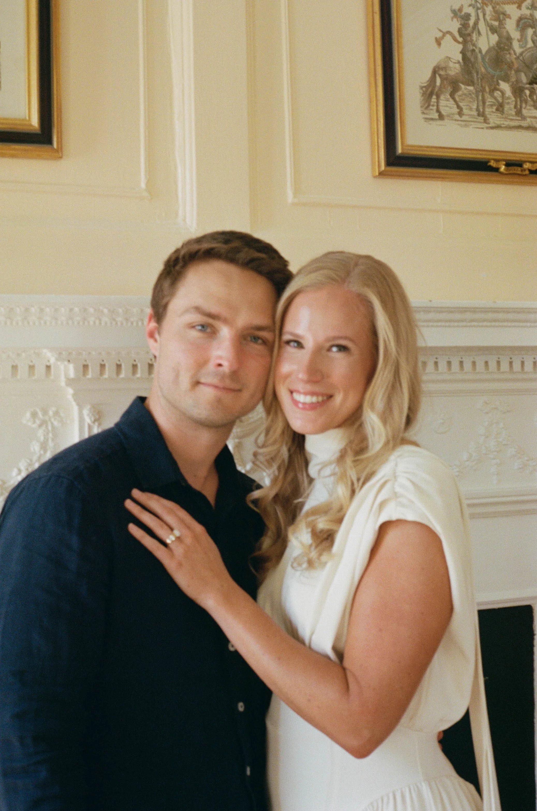 A close-up photo of a smiling couple embracing each other indoors, with the man's arm around the woman's waist. The man has short brown hair and is wearing a dark blue shirt. The woman has long wavy blonde hair and is wearing a white dress with puffe