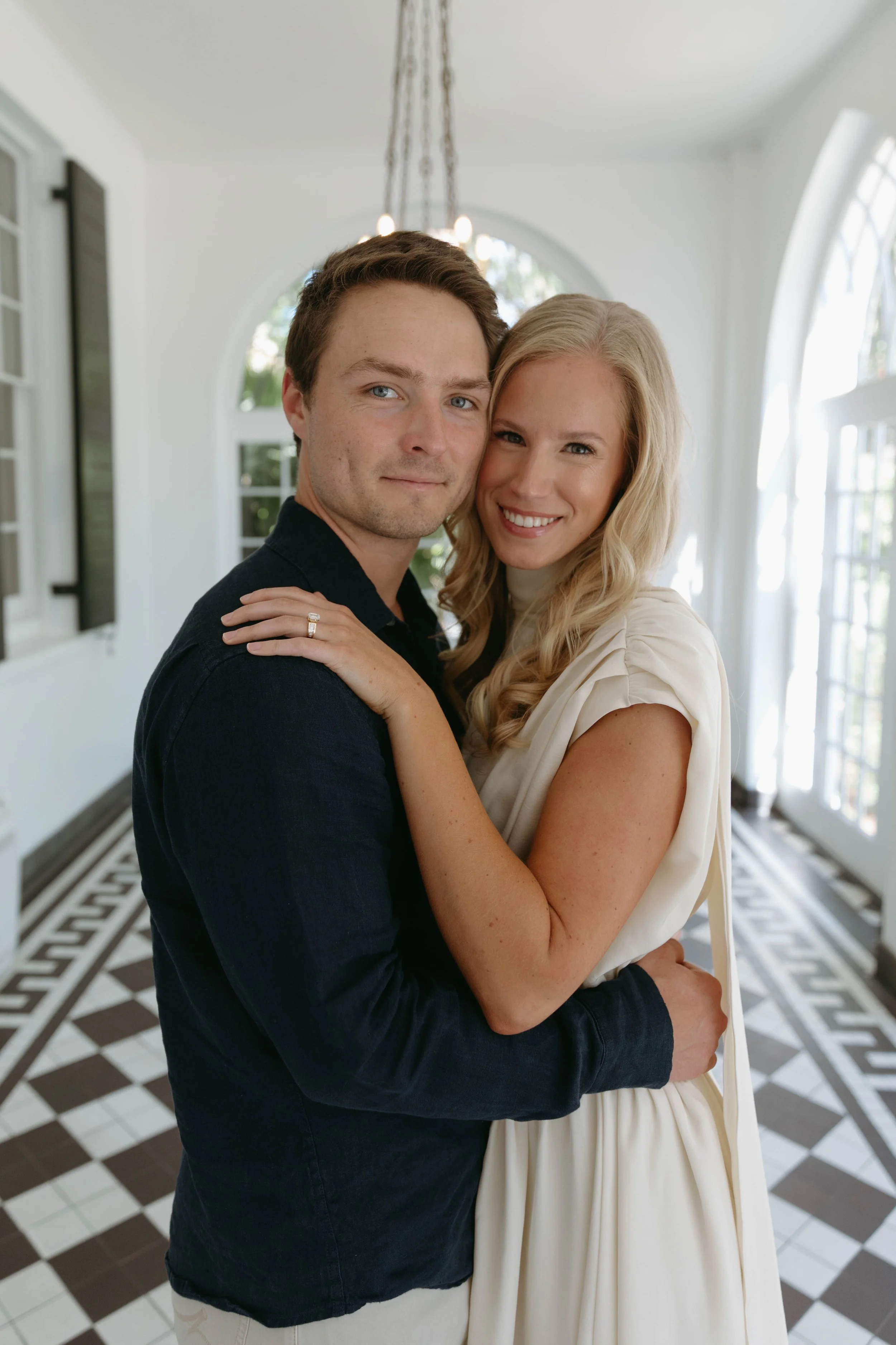 A couple embracing indoors with patterned black and white floor and large windows in the background.