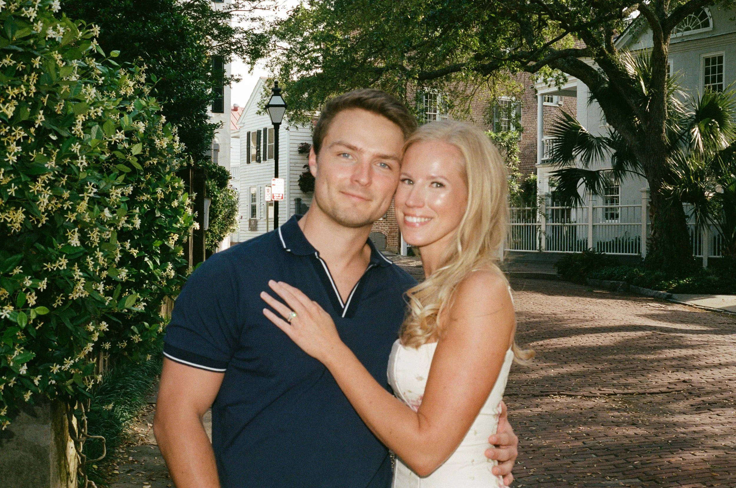 A smiling couple embracing on a brick sidewalk in a residential neighborhood with trees and houses in the background.