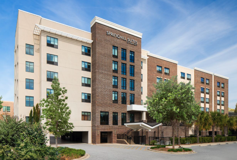 Exterior view of SpringHill Suites by Marriott, a multi-story hotel building with brick and beige facade, surrounded by trees and landscaping, under a blue sky.