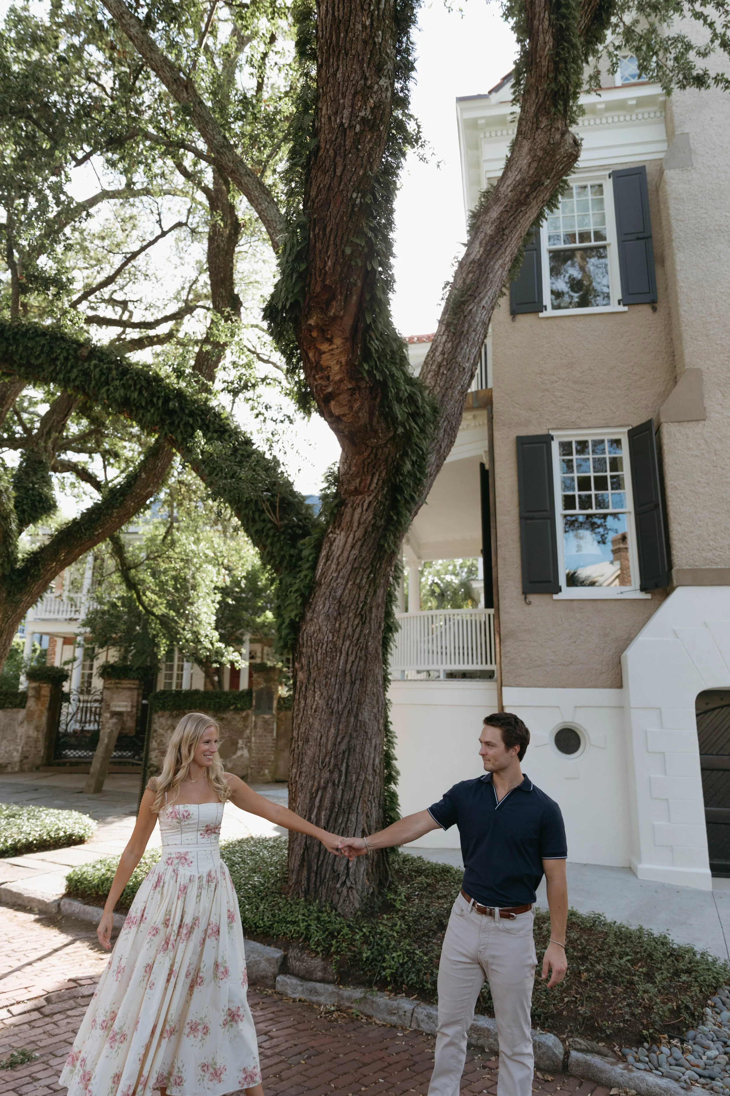 A young woman in a floral dress and a young man in a navy polo shirt holding hands and dancing in a garden with trees and a white house with blue shutters in the background.