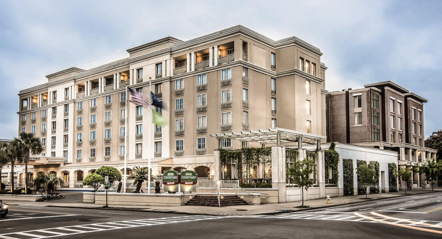 Exterior view of a multi-story Hilton hotel with flags, palm trees, and greenery, located on a city street at dusk.