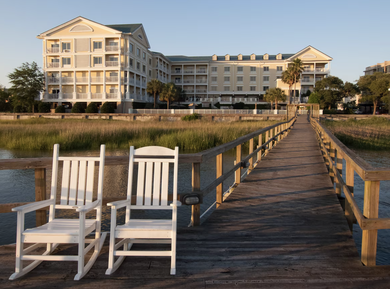 A wooden dock extends over a body of water, with a white rocking chair and a white lawn chair on the dock, leading to a large multi-story white building with balconies, surrounded by trees and greenery during sunset.