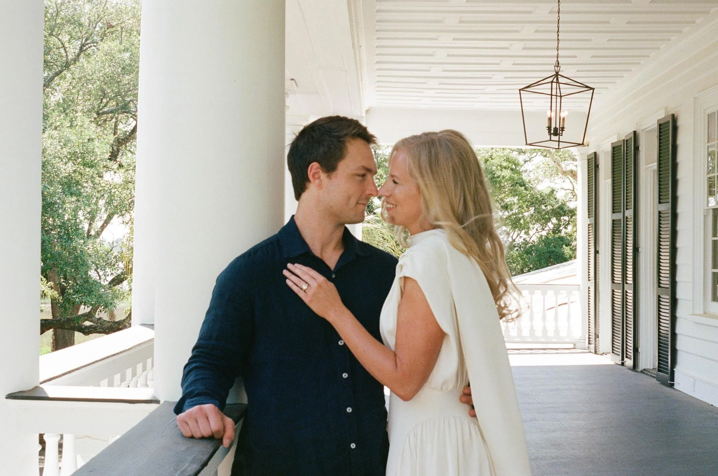 A couple standing close on a porch, facing each other with noses touching. The man has dark hair and is wearing a navy blue shirt. The woman has blonde hair and is wearing a white dress. Green trees are visible in the background.