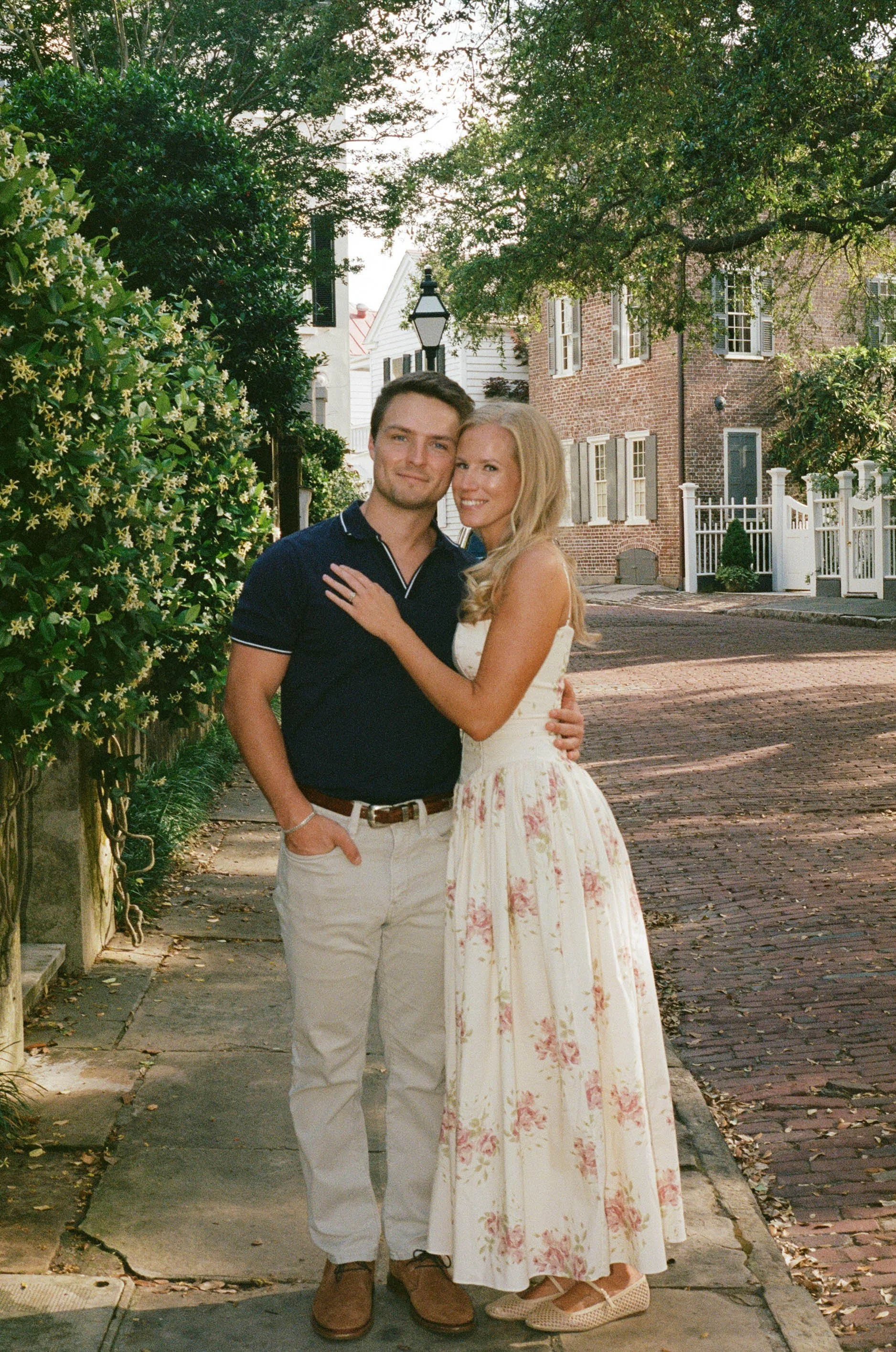 A smiling young couple standing close together outdoors in a quaint neighborhood, with greenery and brick houses in the background. The man is wearing a dark polo shirt and light-colored pants, and the woman is wearing a white floral dress.