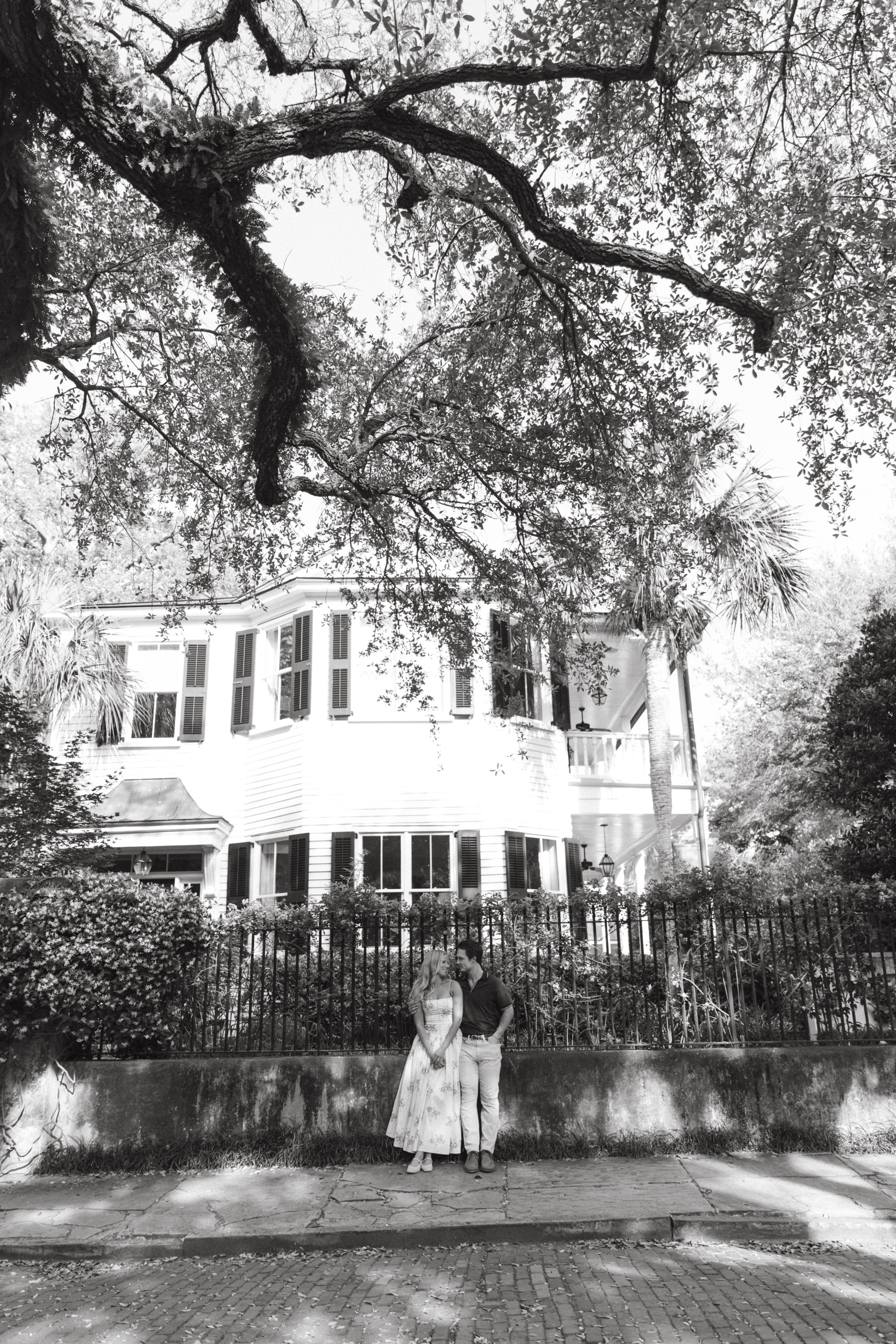 A black-and-white photo of a couple standing in front of a black wrought iron fence, with a large white multi-story house and tall trees in the background. The woman wears a long dress and the man wears a dark shirt and light pants.