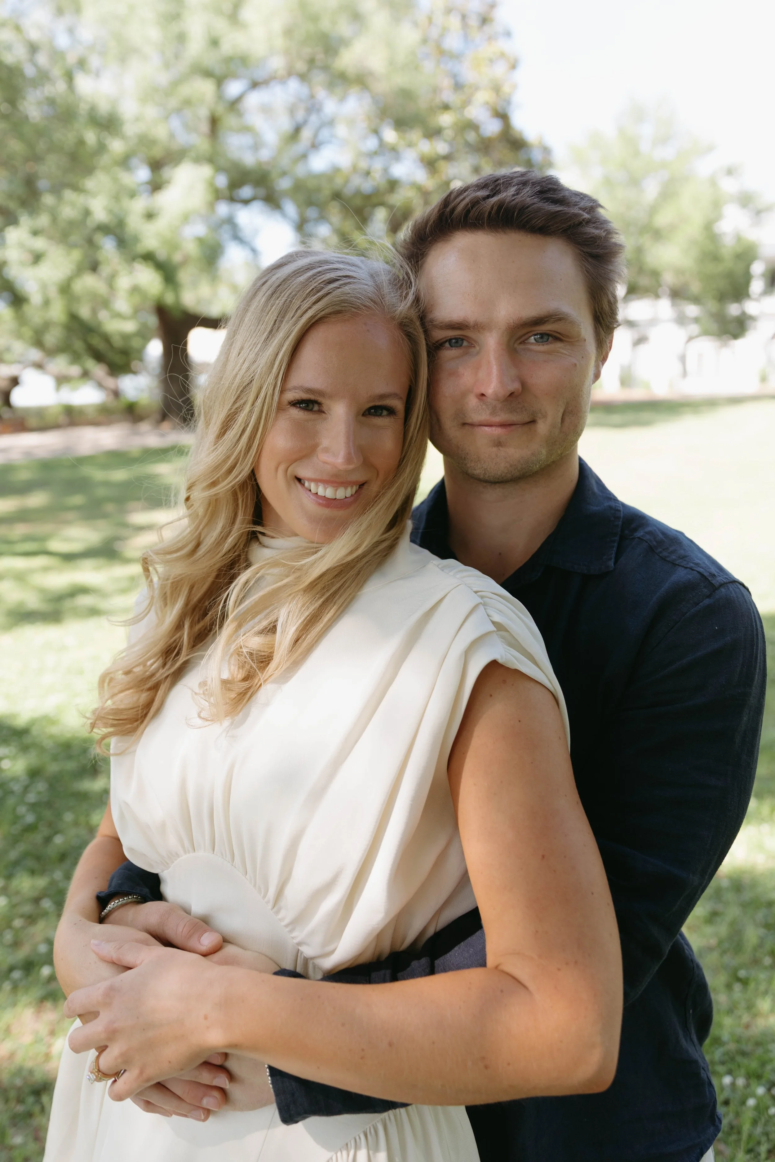 A smiling woman with long blonde hair and a man with short dark hair, both embracing outdoors on a sunny day with green trees in the background.