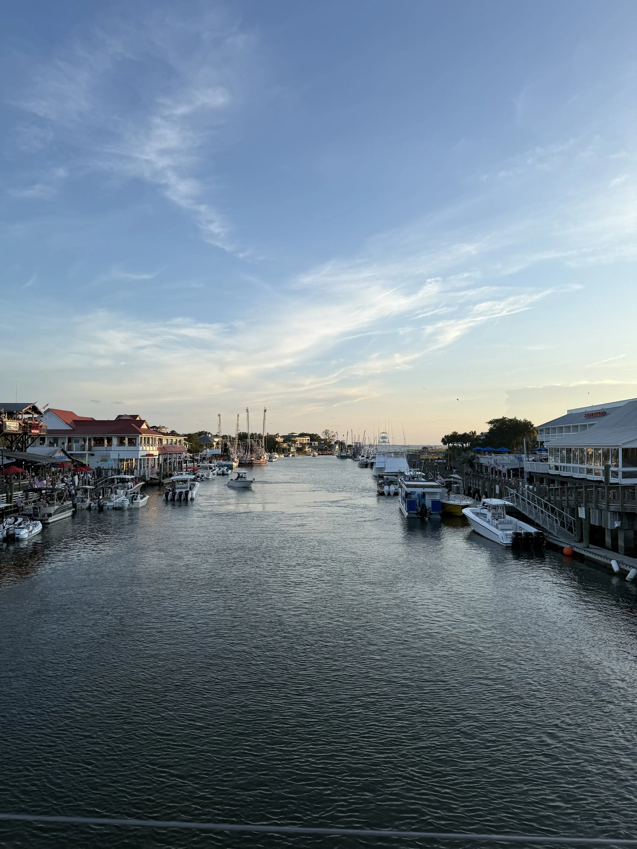 A marina with boats docked along the waterfront, buildings on either side, and a sky with scattered clouds at sunset.