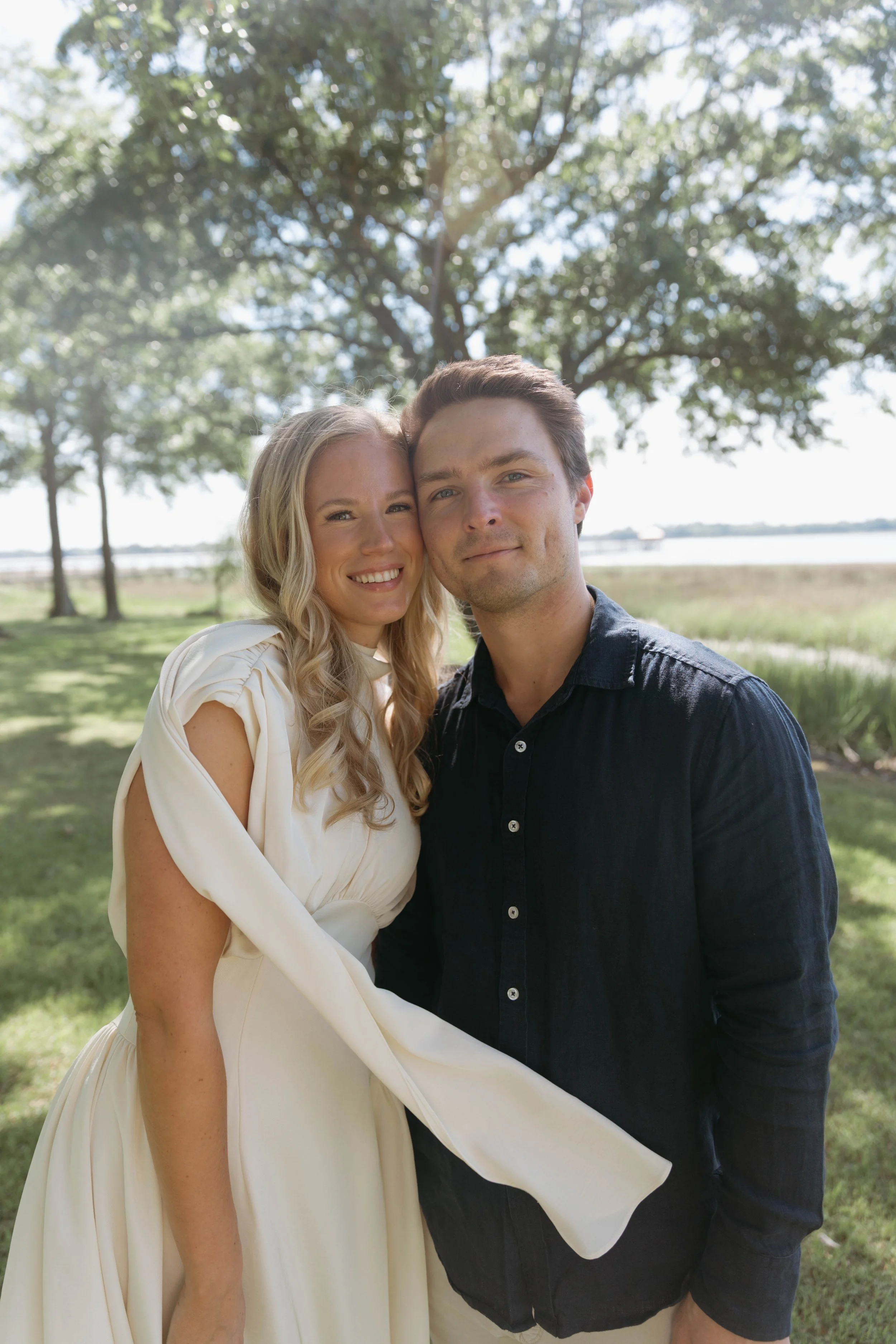 A smiling blonde woman in a cream dress standing next to a smiling man in a dark shirt. They are outdoors under a large tree.