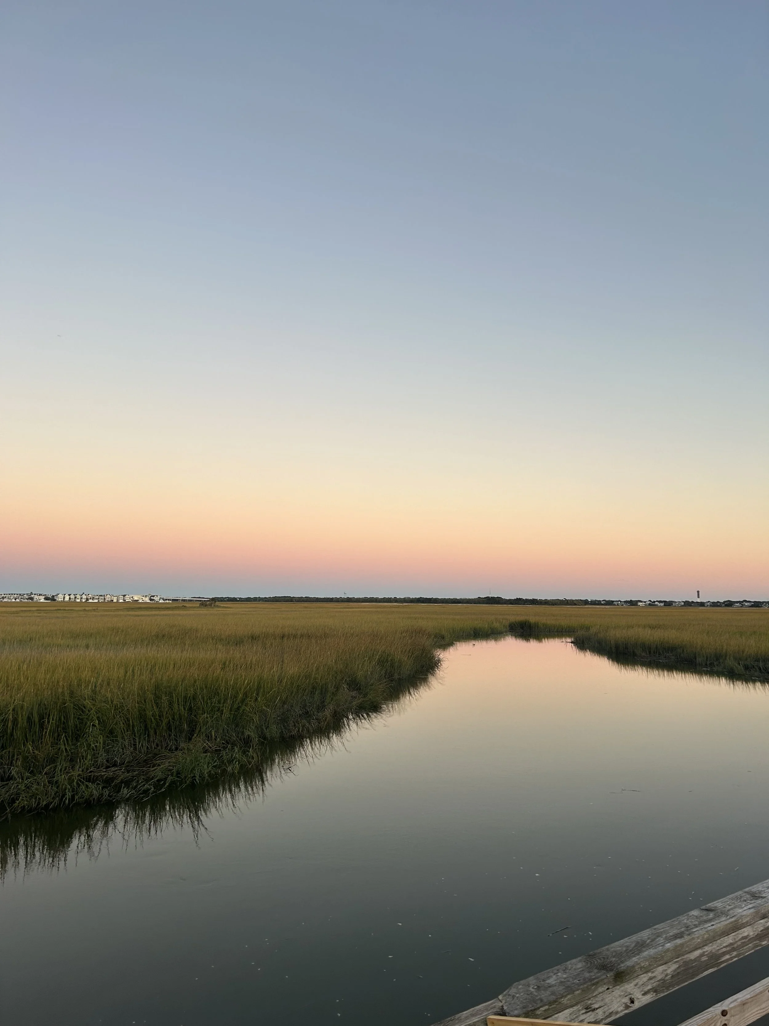 A calm waterway reflecting the pastel-colored sky at sunset, surrounded by green marsh grass, with a wooden dock or pier in the foreground and a distant horizon with buildings and a tower.