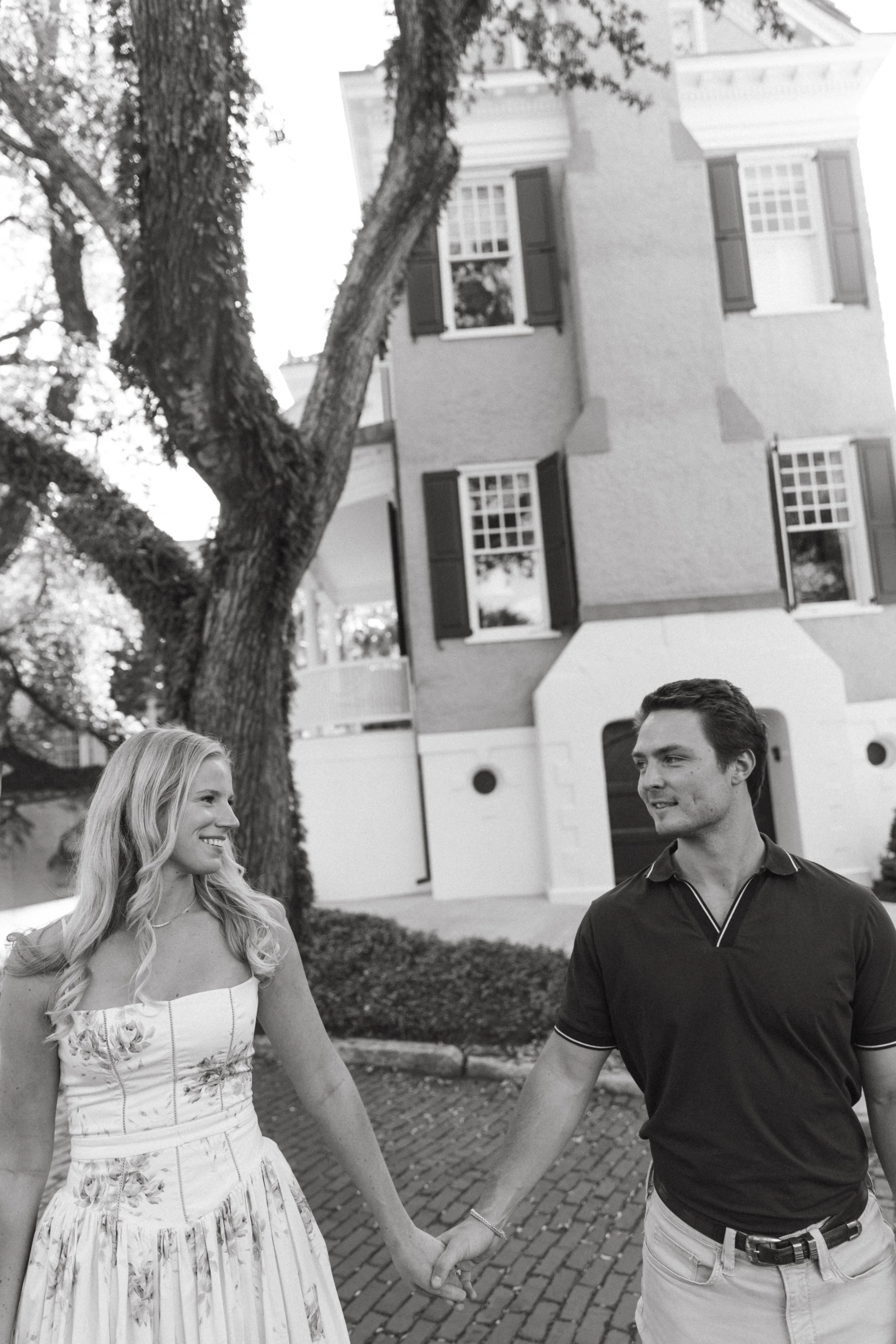 A couple is holding hands and looking at each other outside in a park or garden area, with a residential building and large trees in the background.