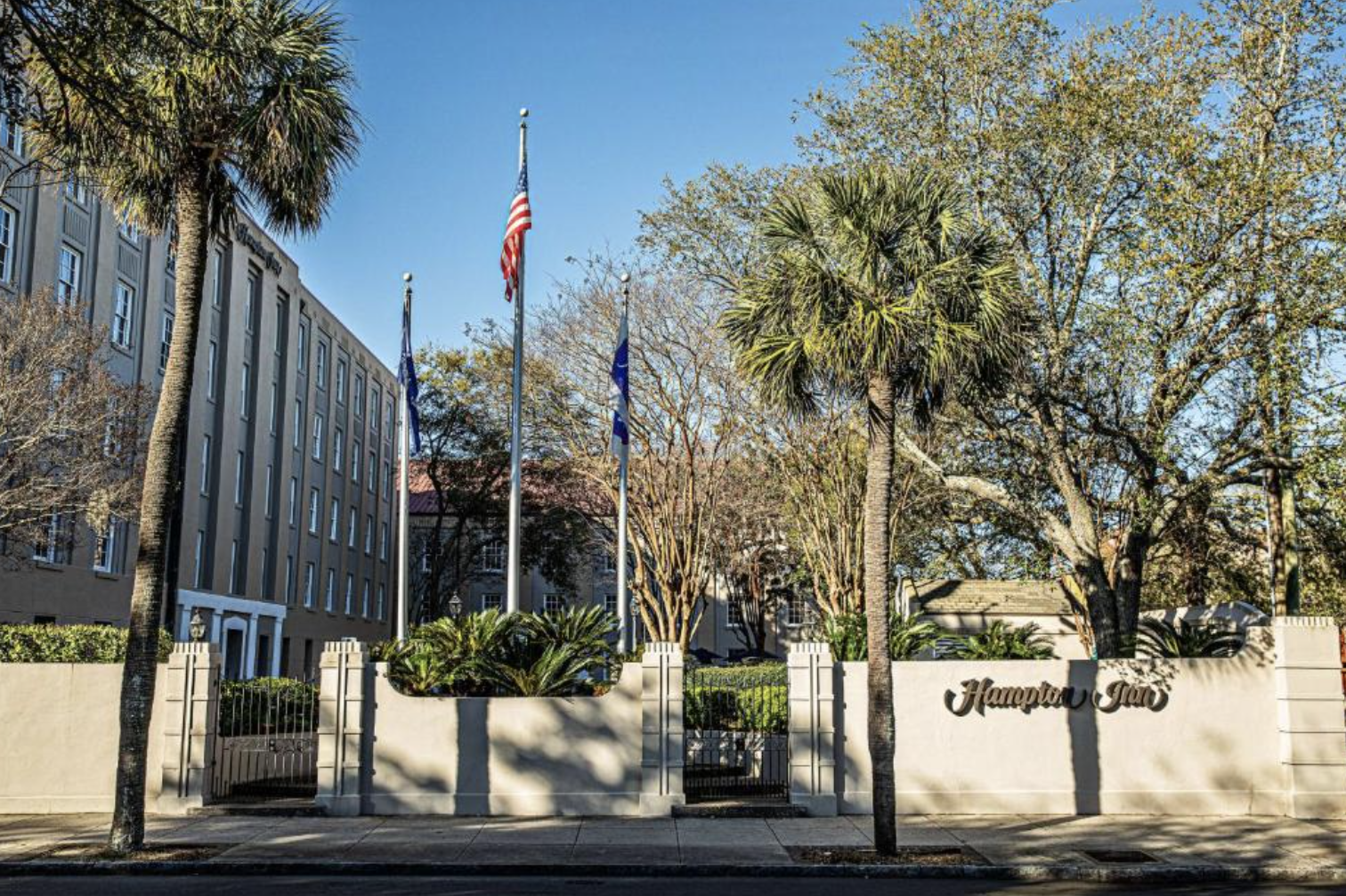 Entrance to Hampton Fay with a white wall, black gate, and well-maintained garden, two palm trees, several other trees, and American, Nevada, and another unidentified flag flying on poles. A multi-story building is visible to the left in the background.