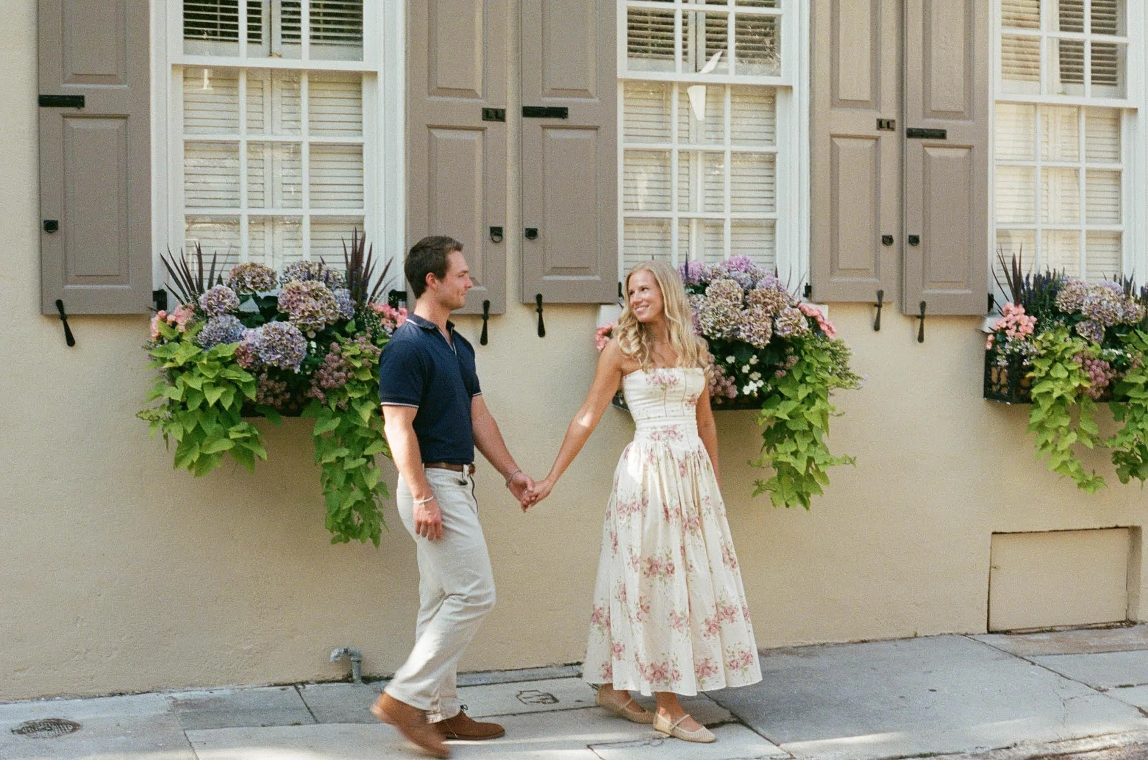 A young couple holding hands and smiling in front of a beige house with gray shutters and purple floral window boxes.