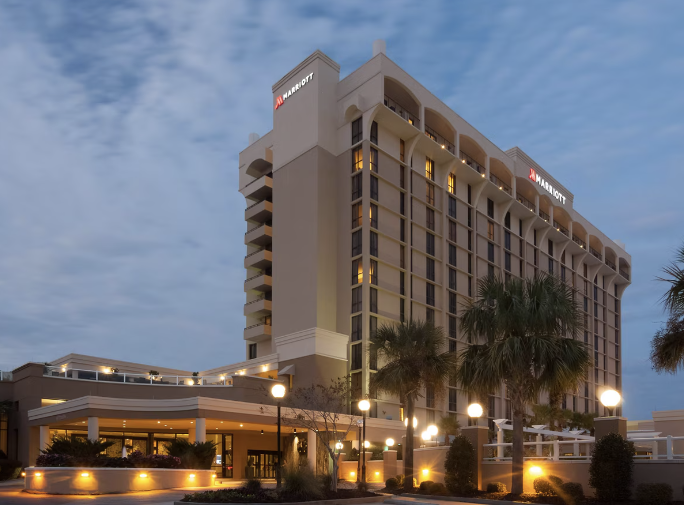 A tall Marriott hotel building with illuminated windows and palm trees in the foreground, taken at dusk.