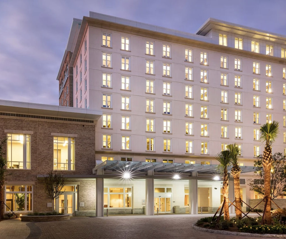 A modern multi-story hotel building with lit windows at dusk, palm trees in the foreground, and a covered entrance.